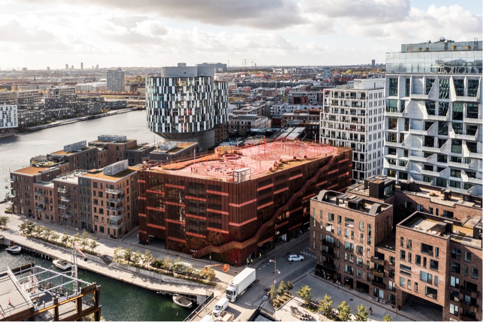 Städtische Architektur mit modernen Wohn- und Bürogebäuden entlang eines Kanals, darunter ein auffälliges rotes Gebäude mit Dachterrasse und ein rundes Gebäude mit gemusterten Fassaden.