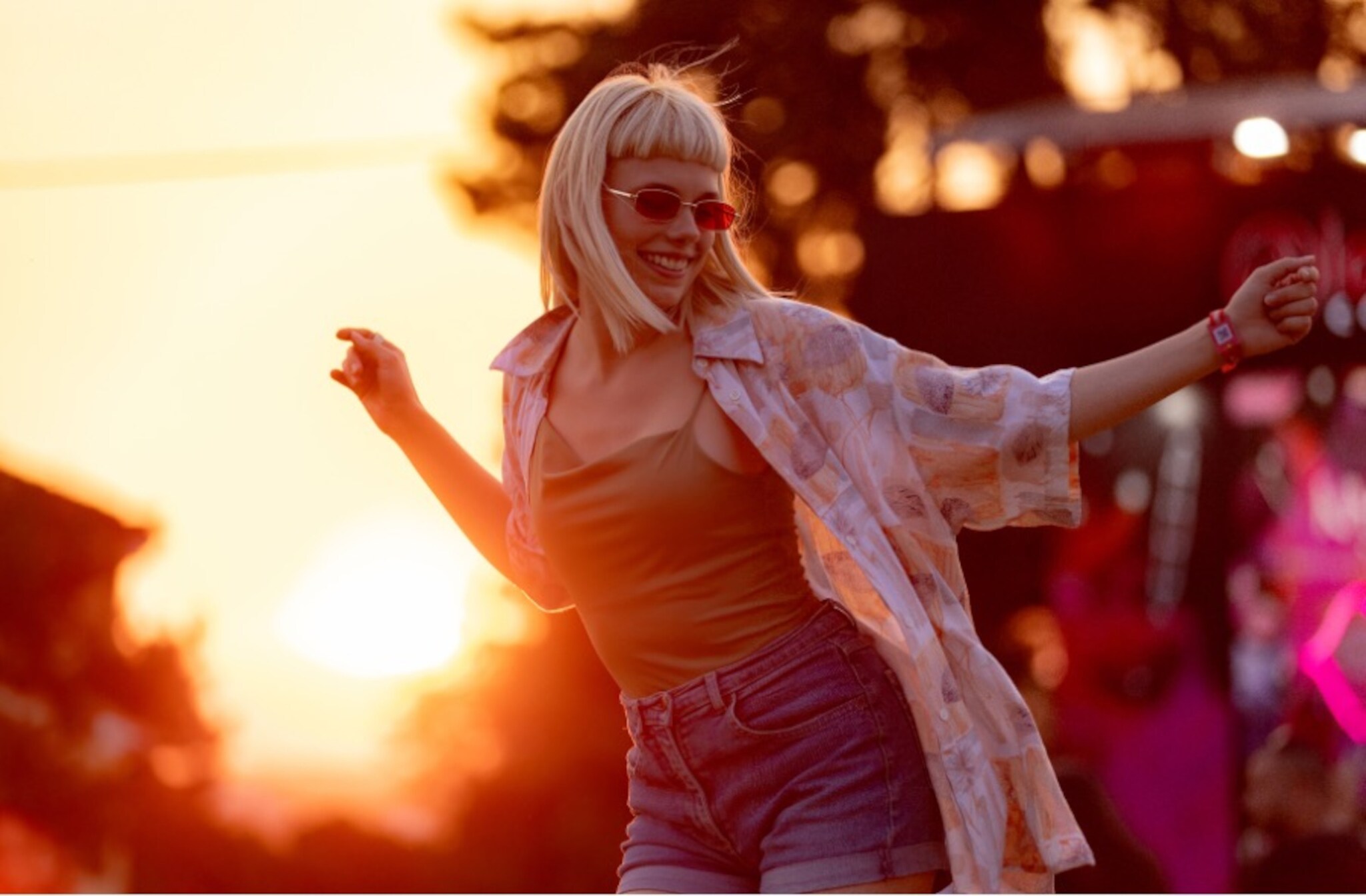 Eine stylisch gekleidete Frau in Shorts mit Sonnenbrille und kurzem Pony tanzt auf einem Festivalgelände im warmen Licht bei Sonnenuntergang.