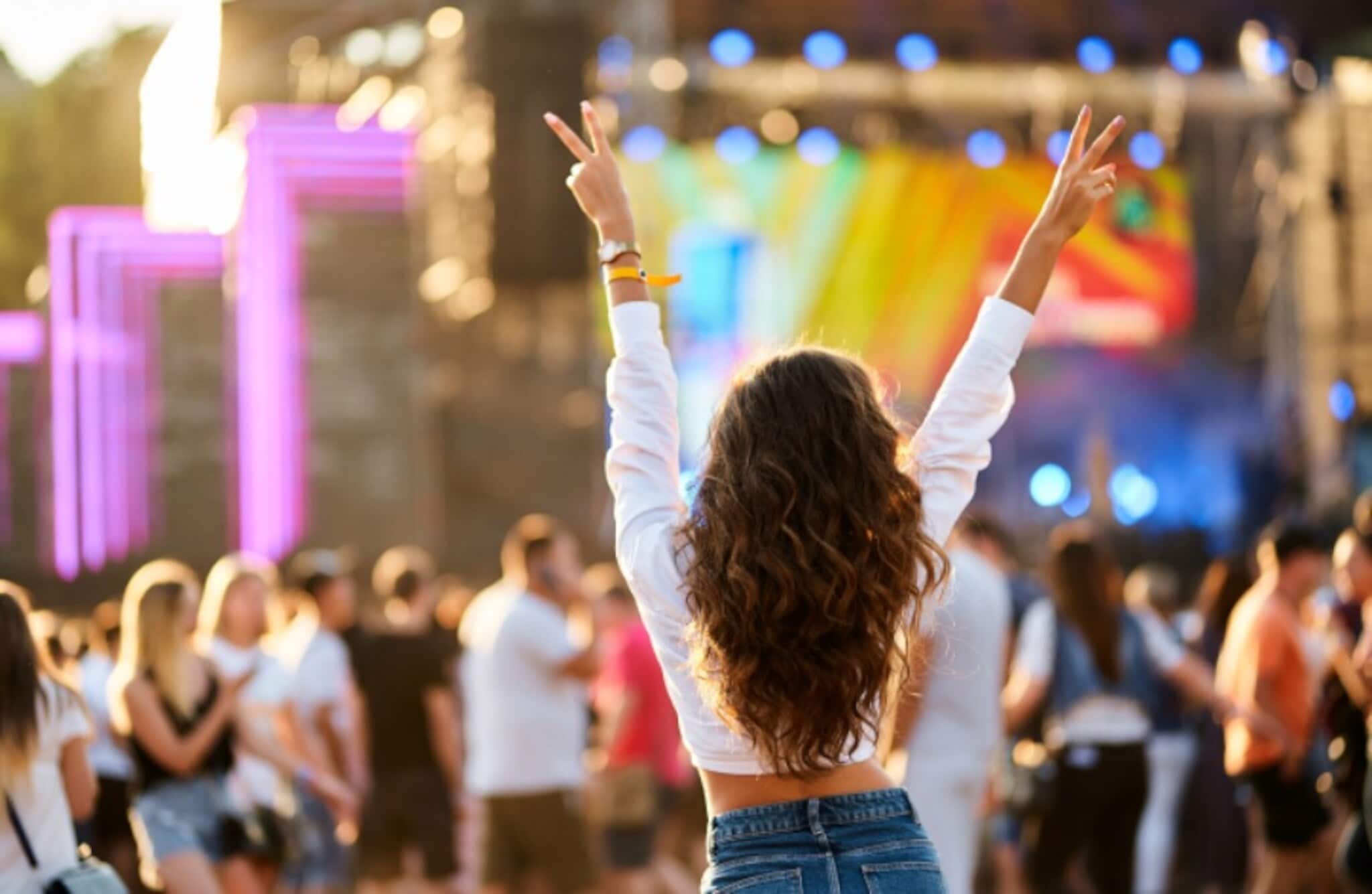Rückansicht einer jungen Frau, die ihr Hände mit Peace-Zeichen in die Luft hebt vor einer Musikbühne mit Publikum im Freien. Rückansicht einer jungen Frau, die ihr Hände mit Peace-Zeichen in die Luft hebt vor einer Musikbühne mit Publikum im Freien.
