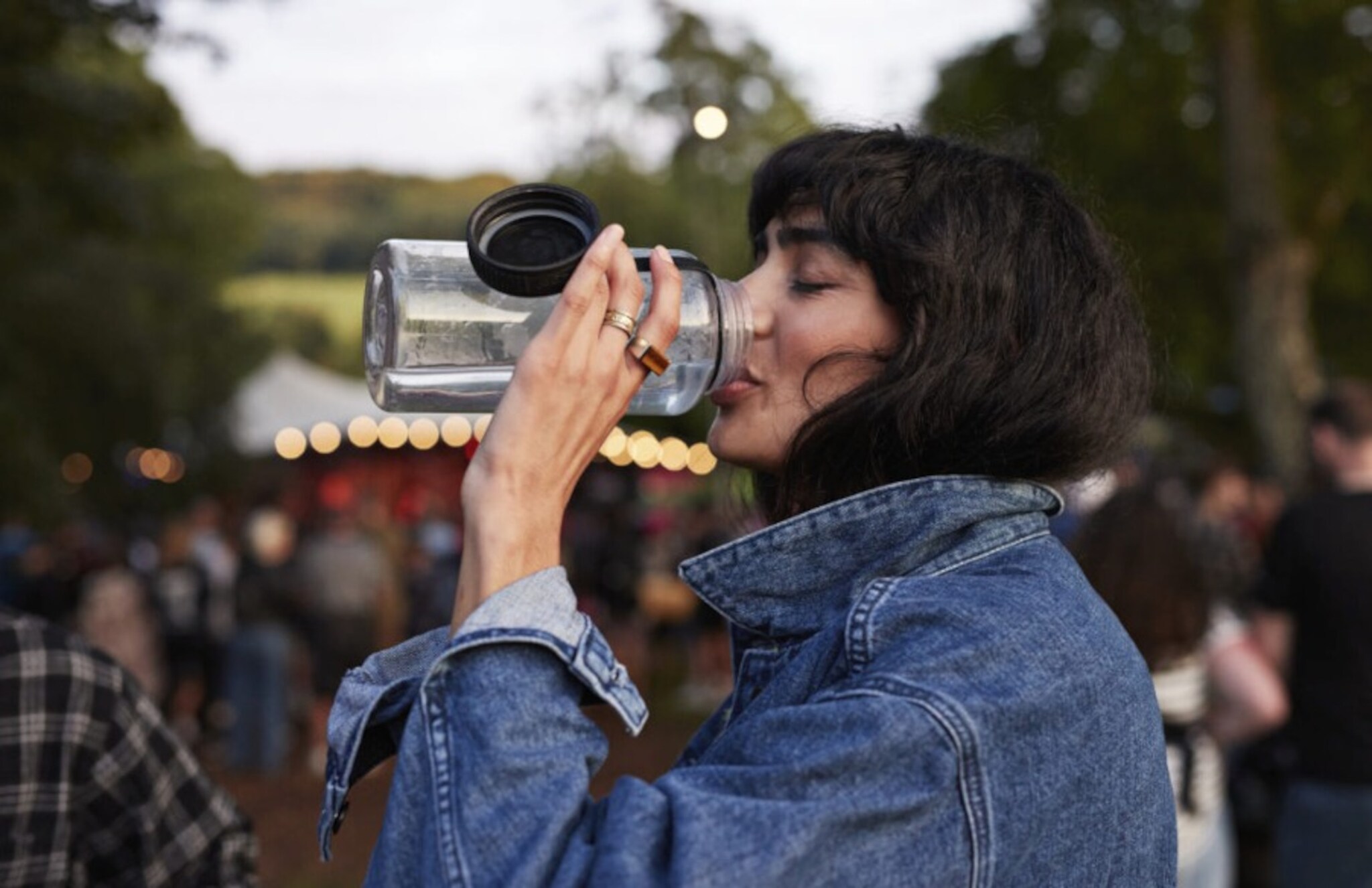 Eine junge Frau in Jeansjacke trinkt aus einer großen Wasserflasche auf einem Festivalgelände.