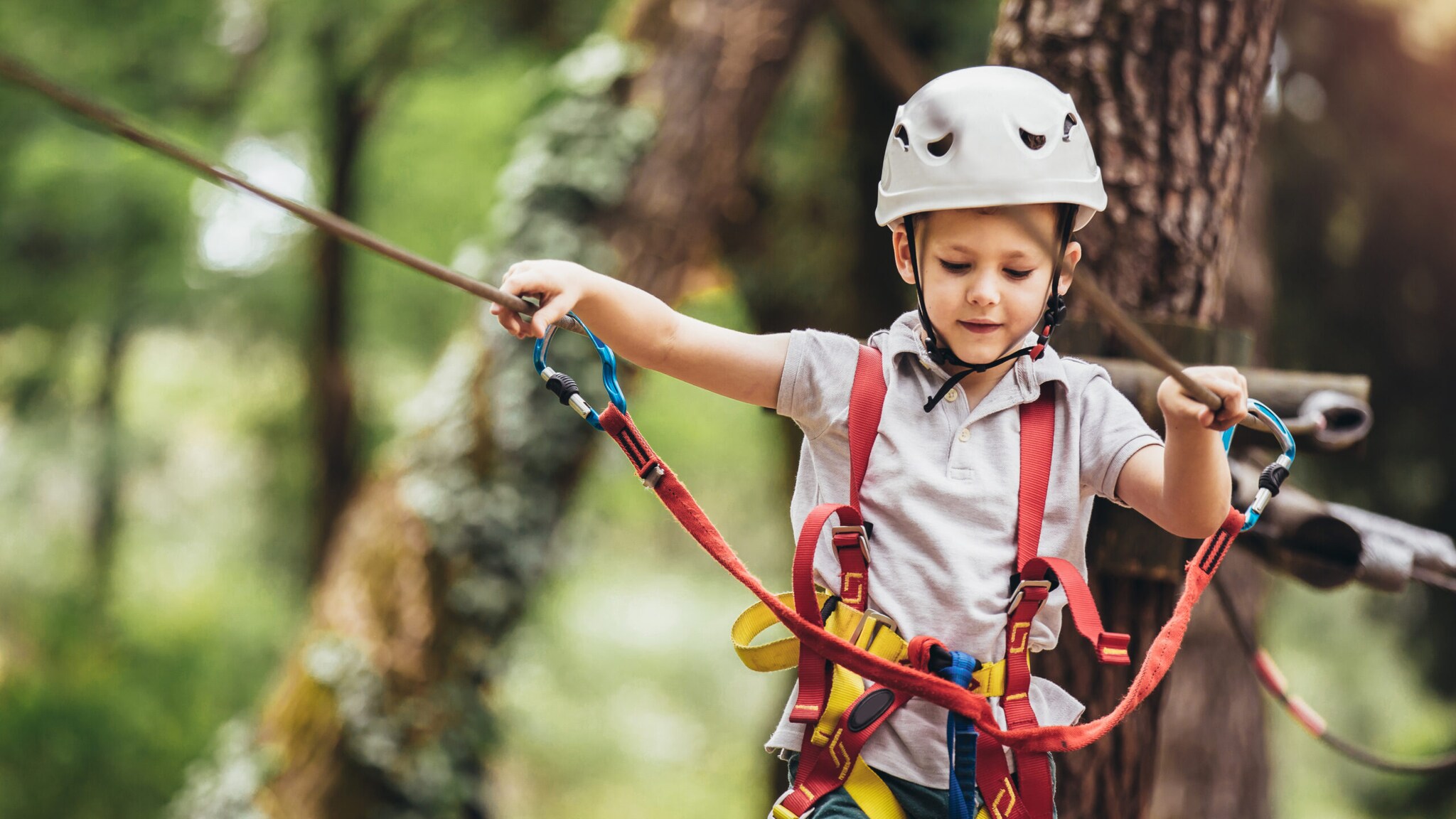 Kind mit weißem Helm und Kletterausrüstung hält sich an Seilen in einem Hochseilgarten fest