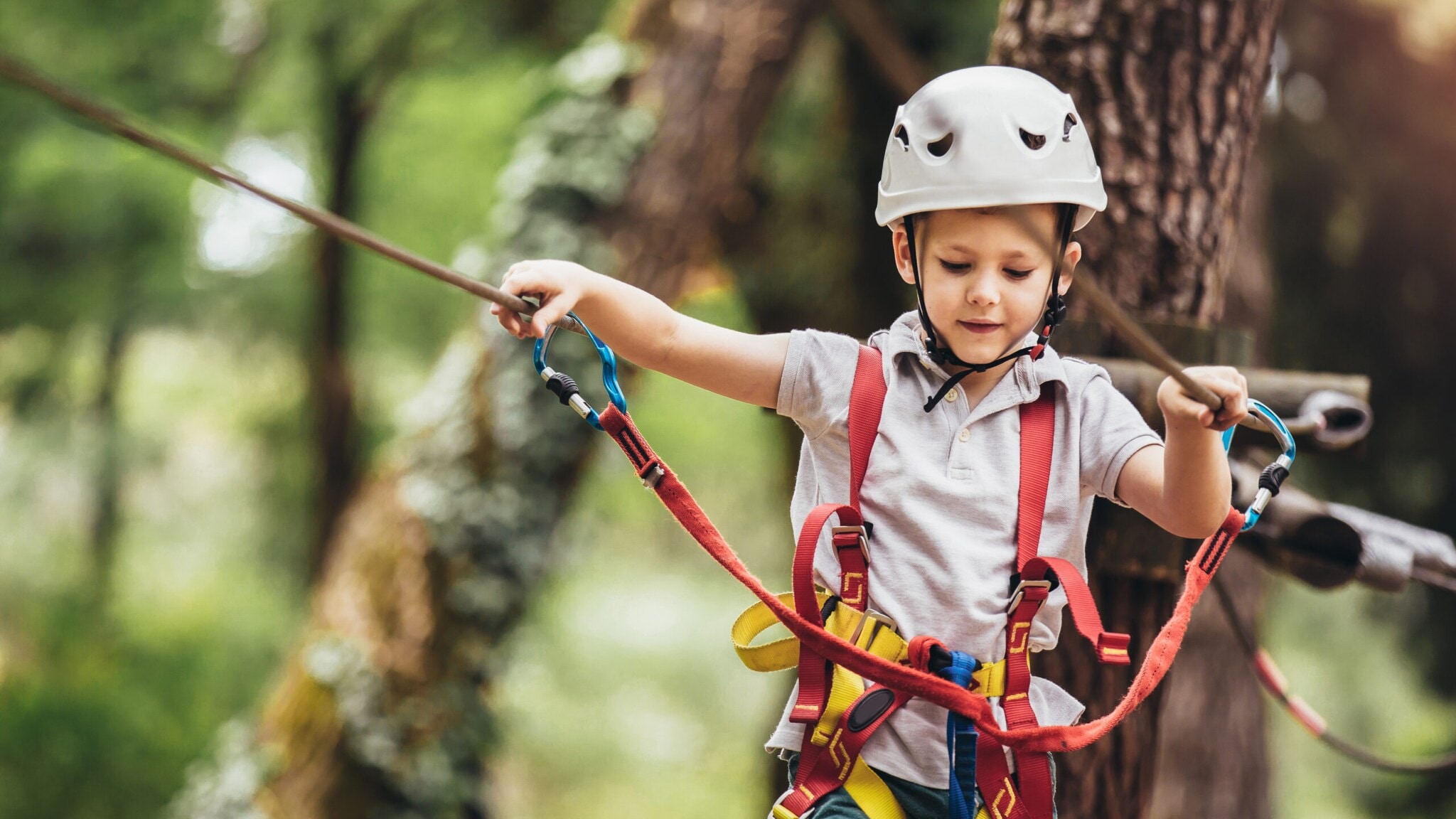 Kind mit weißem Helm und Kletterausrüstung hält sich an Seilen in einem Hochseilgarten fest Kind mit weißem Helm und Kletterausrüstung hält sich an Seilen in einem Hochseilgarten fest