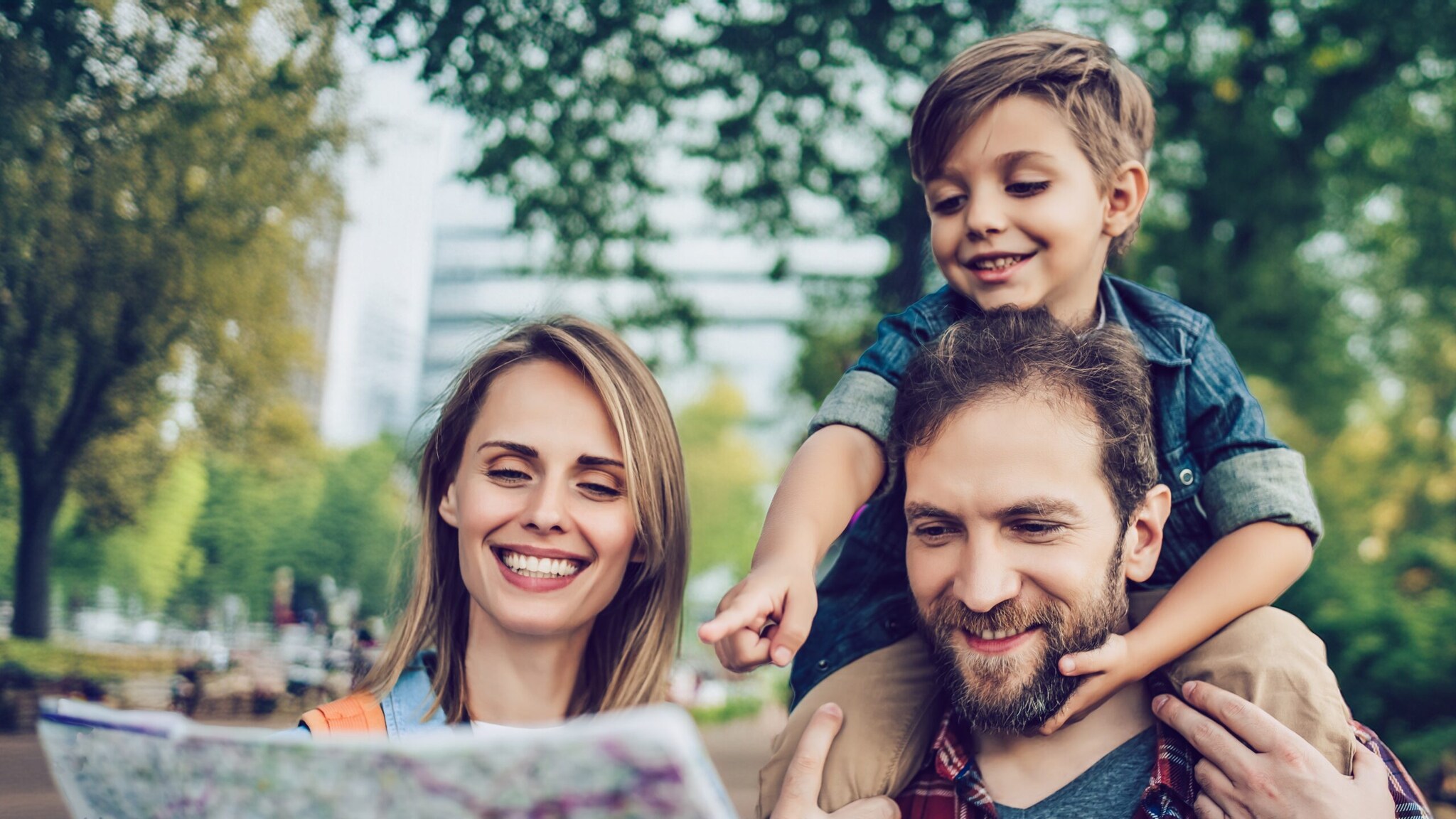 Vater trägt Sohn auf Schultern, Mutter hält Karte, sie stehen in einem Park mit Bäumen und Gebäuden im Hintergrund