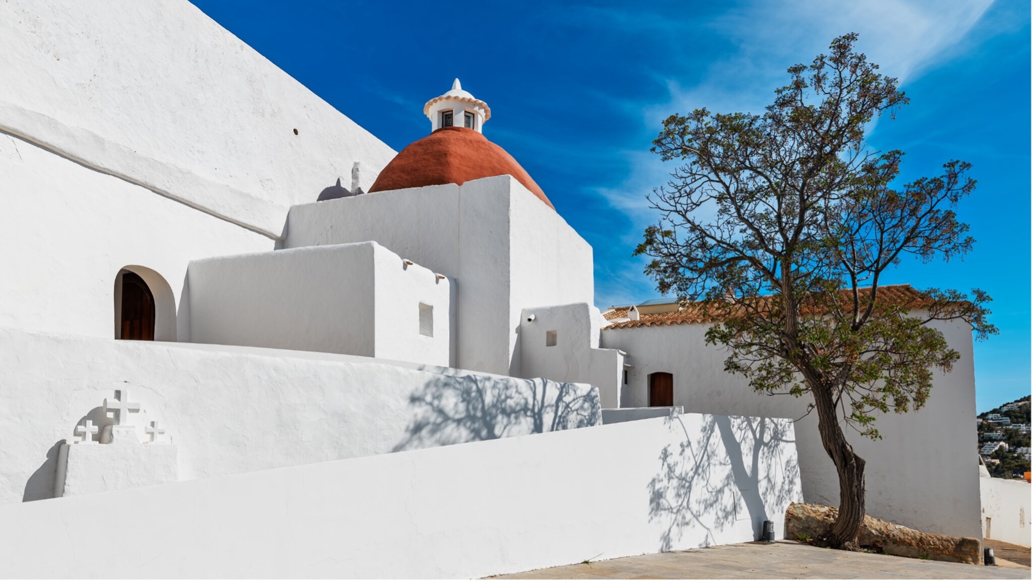 Weiße Gebäude mit roter Kuppel und Baum vor blauem Himmel in mediterraner Landschaft