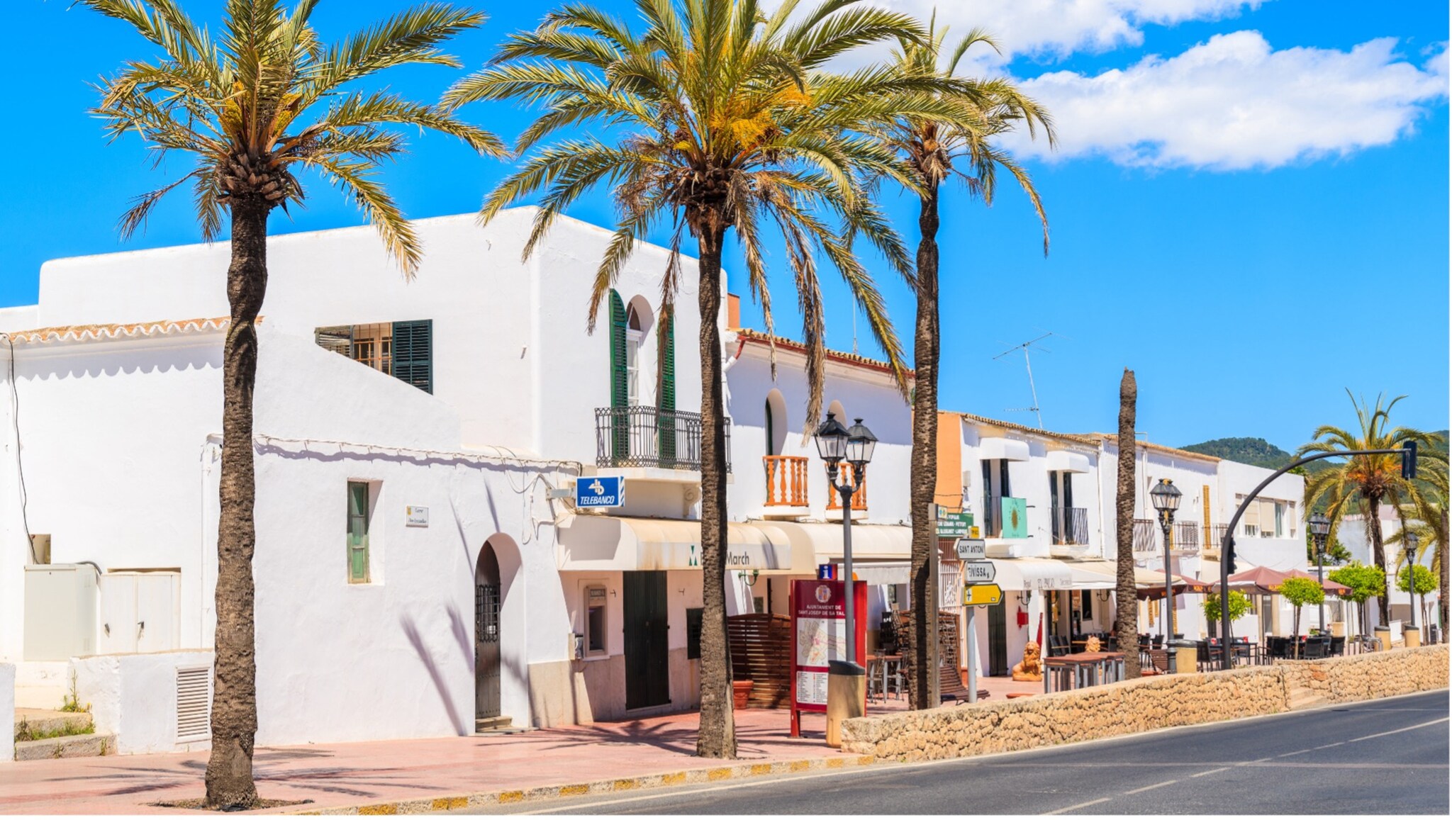 Weiße Gebäude mit Arkaden und Balkonen an einer sonnigen Straße mit Palmen unter blauem Himmel