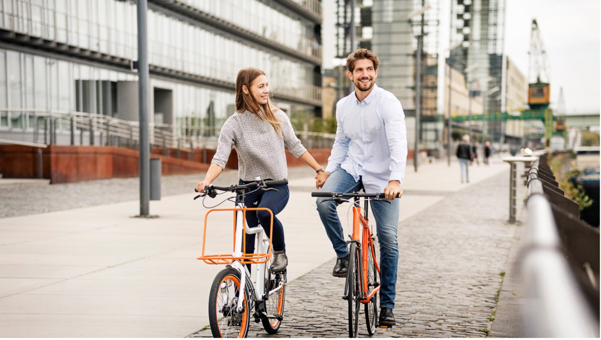 Paar hält Händchen beim Fahrradfahren auf einem gepflasterten Weg in einem modernen Stadtviertel