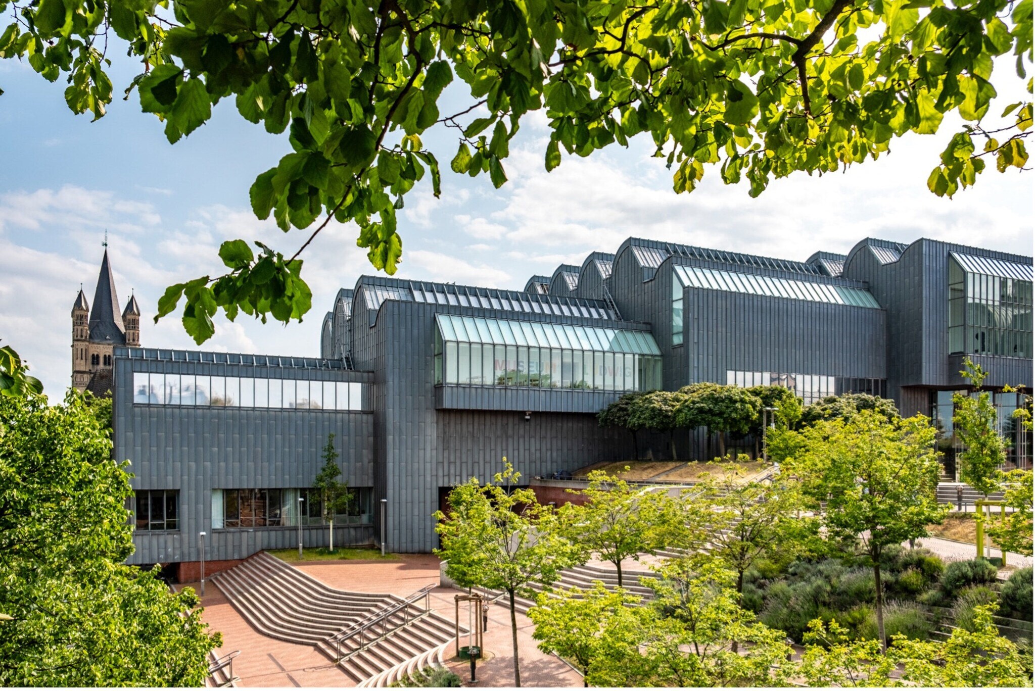 Modernes, mehrteiliges Gebäude mit Glasdächern und Metallfassade, umgeben von Bäumen und Grünflächen, im Hintergrund Kirchturm Modernes, mehrteiliges Gebäude mit Glasdächern und Metallfassade, umgeben von Bäumen und Grünflächen, im Hintergrund Kirchturm