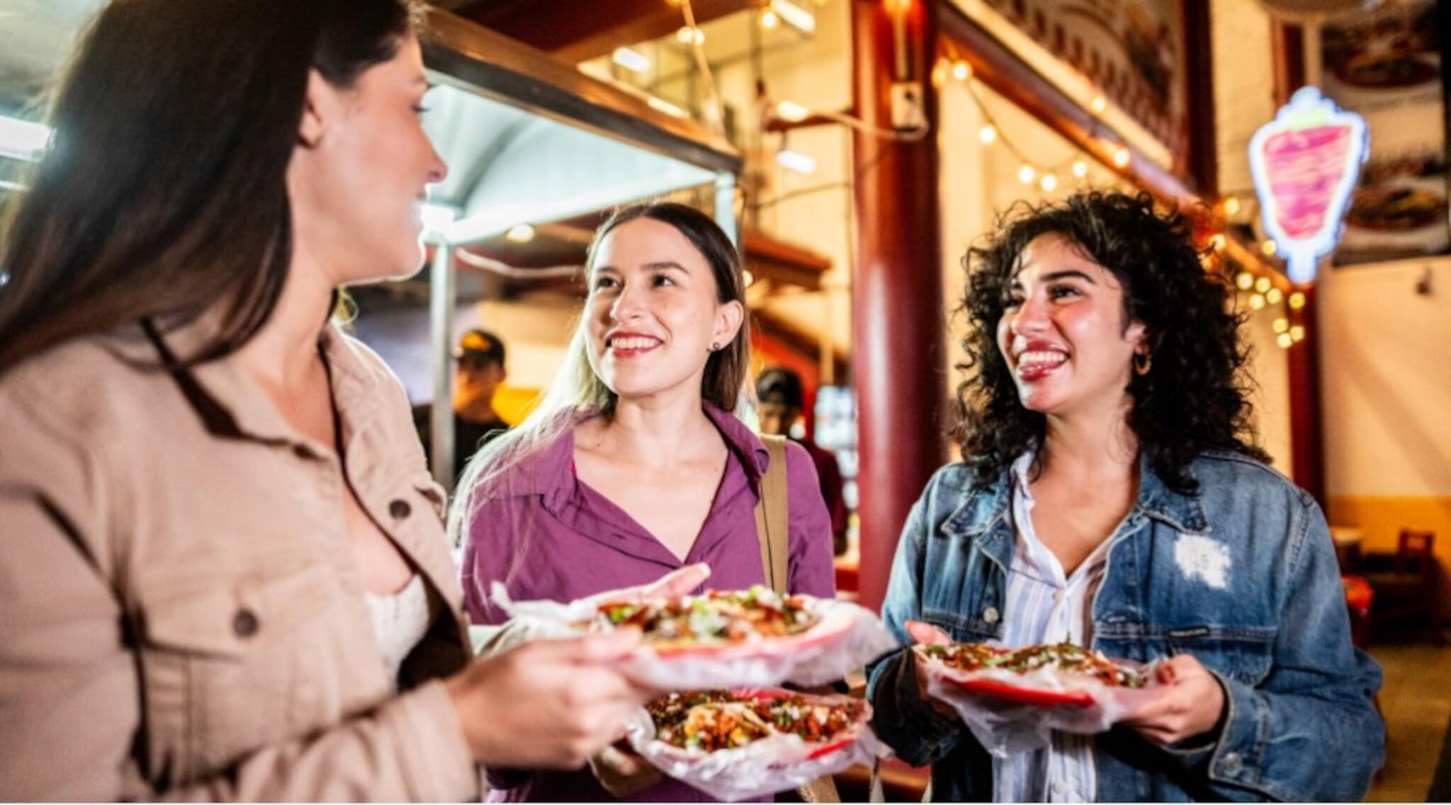 Drei junge Frauen mit Taco-Gerichten in den Händen auf einem beleuchteten Street-Food-Markt bei Nacht. Drei junge Frauen mit Taco-Gerichten in den Händen auf einem beleuchteten Street-Food-Markt bei Nacht.