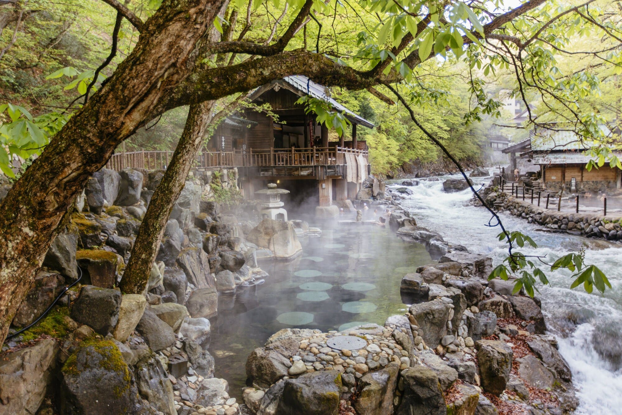 Thermalbad in einem Felsen an einem Fluss mit Holzhütten, umgeben von grünen Bäumen. Thermalbad in einem Felsen an einem Fluss mit Holzhütten, umgeben von grünen Bäumen.