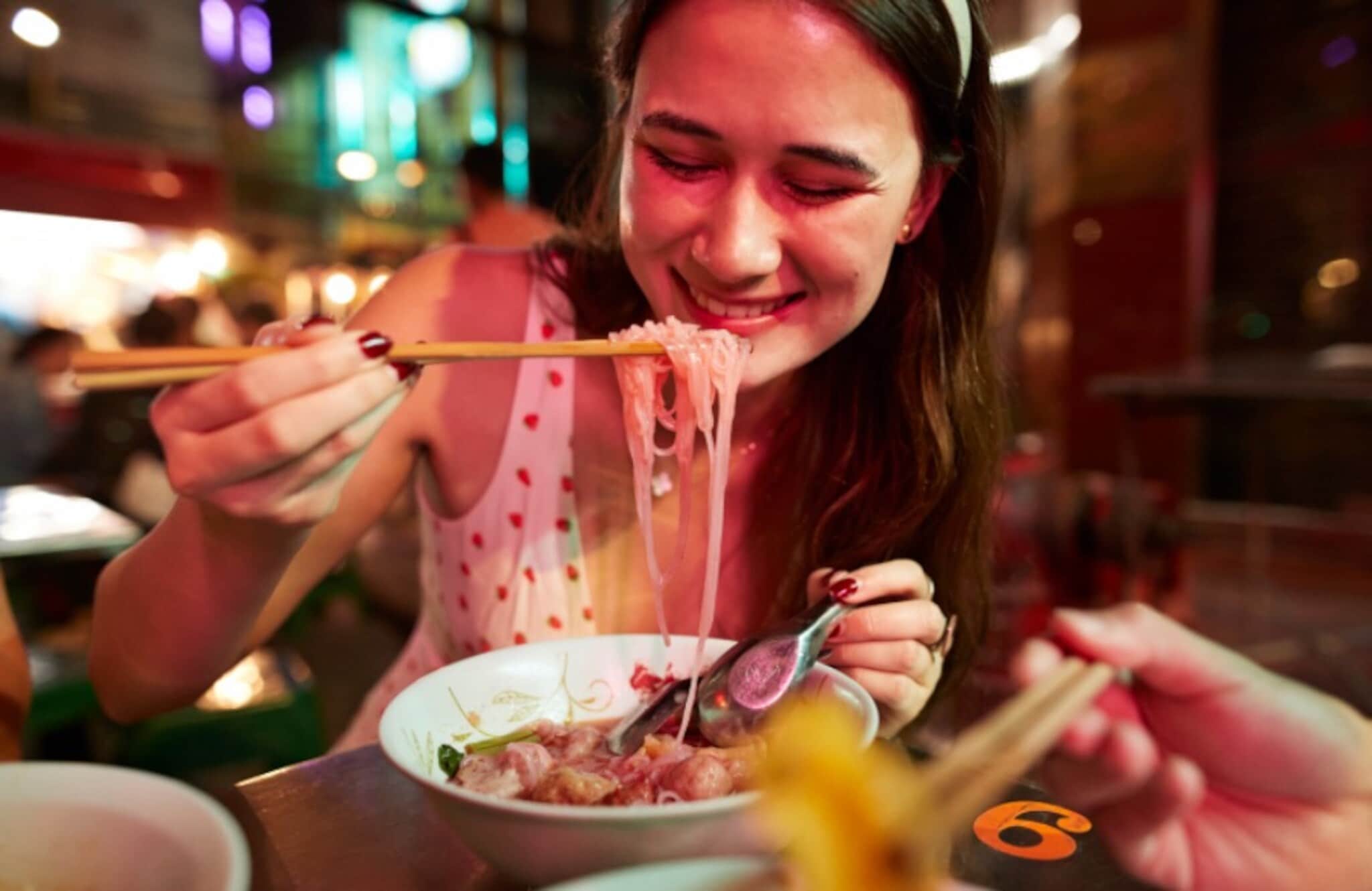 Nahaufnahme einer jungen, lächelnden Frau beim Essen eines asiatischen Nudelgerichtes mit Essstäbchen auf einem Straßenmarkt bei Nacht. Nahaufnahme einer jungen, lächelnden Frau beim Essen eines asiatischen Nudelgerichtes mit Essstäbchen auf einem Straßenmarkt bei Nacht.