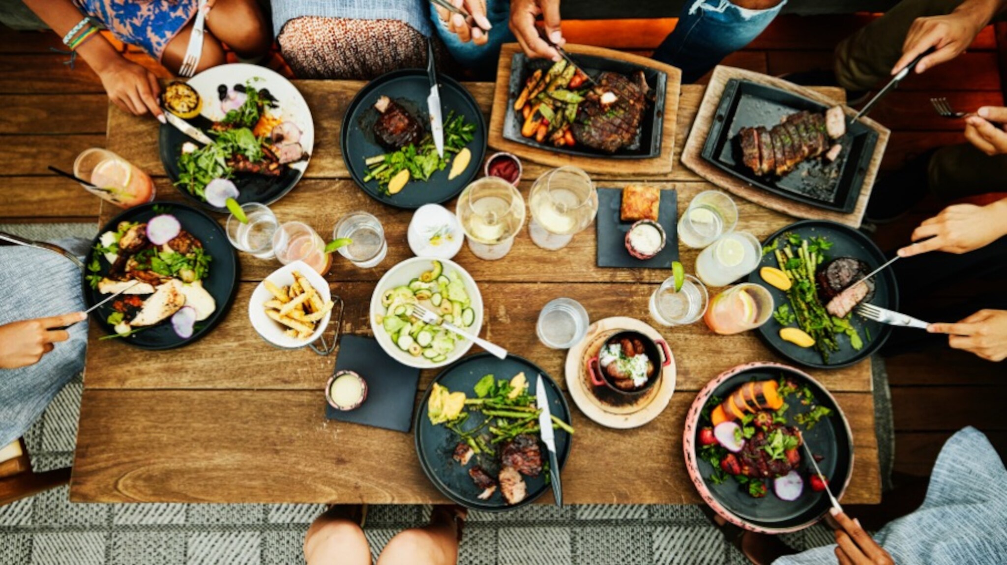 Aufsicht eines Holztisches mit unterschiedlichen Fleischgerichten und Getränken, am Rande die Hände mehrerer Personen mit Besteck.