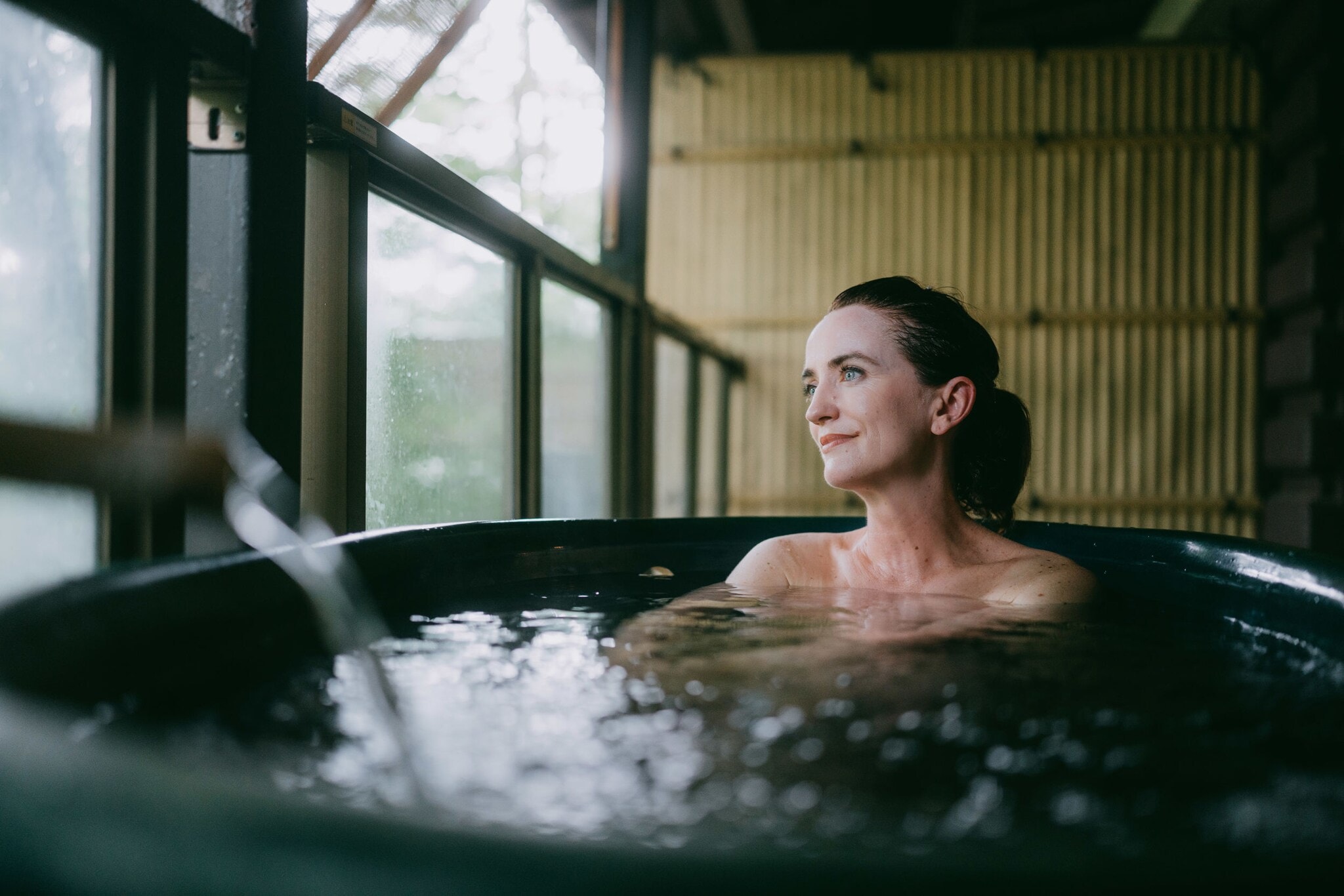 Eine Frau sitzt in einem Thermalbad aus dunklem Stein in einem japanischen Spa mit Bambuswand und schaut zufrieden durch ein Fenster hinaus. Eine Frau sitzt in einem Thermalbad aus dunklem Stein in einem japanischen Spa mit Bambuswand und schaut zufrieden durch ein Fenster hinaus.
