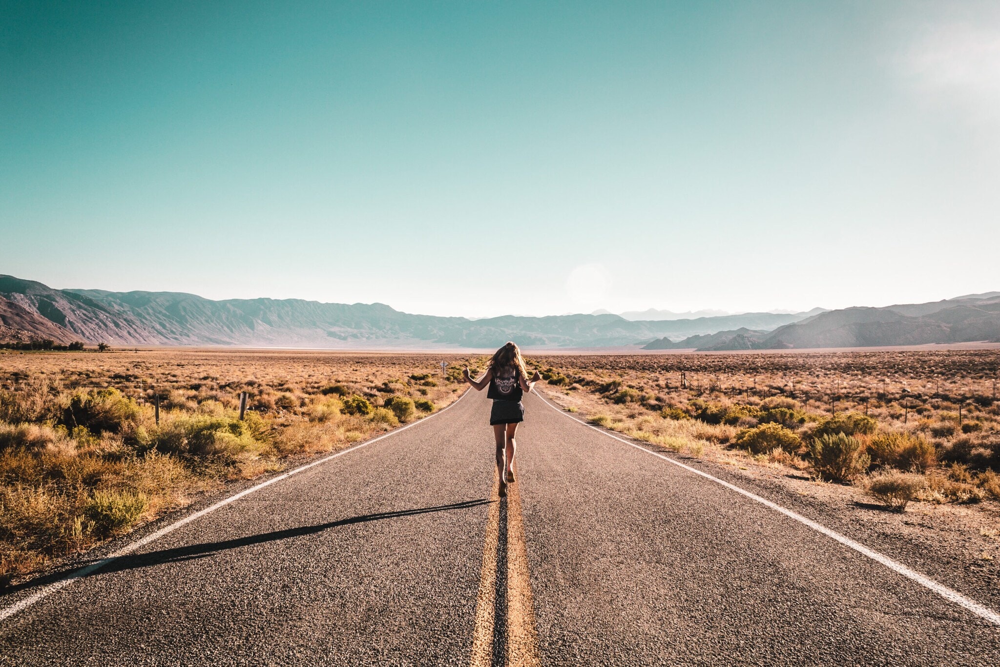 Eine Frau mit Rucksack läuft mitten auf einer leeren Straße, im Hintergrund sind Berge. Eine Frau mit Rucksack läuft mitten auf einer leeren Straße, im Hintergrund sind Berge.