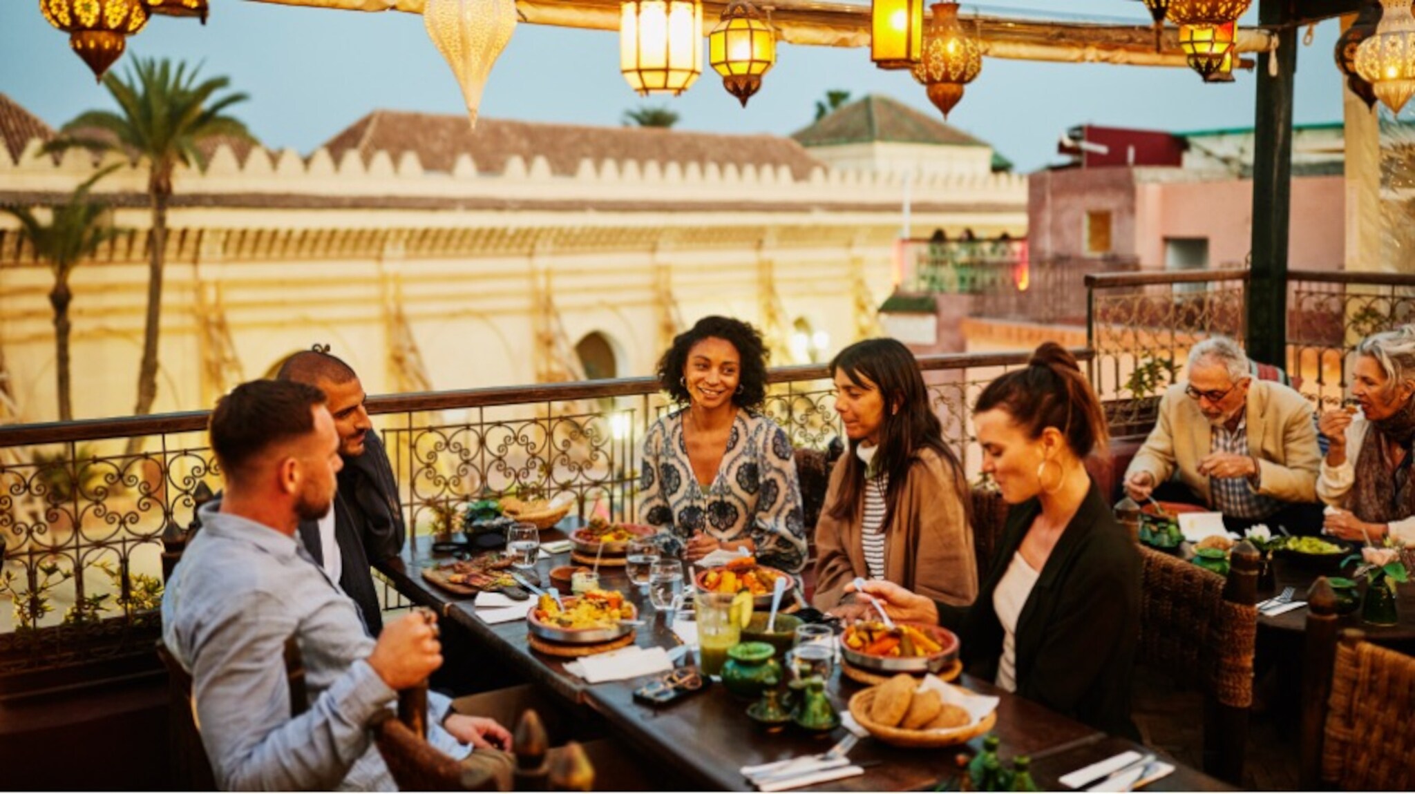 Eine Personengruppe an einem Tisch beim Essen auf einer mit orientalischen Lampen geschmückten Dachterrasse in einem Stadtzentrum mit arabischer Architektur.