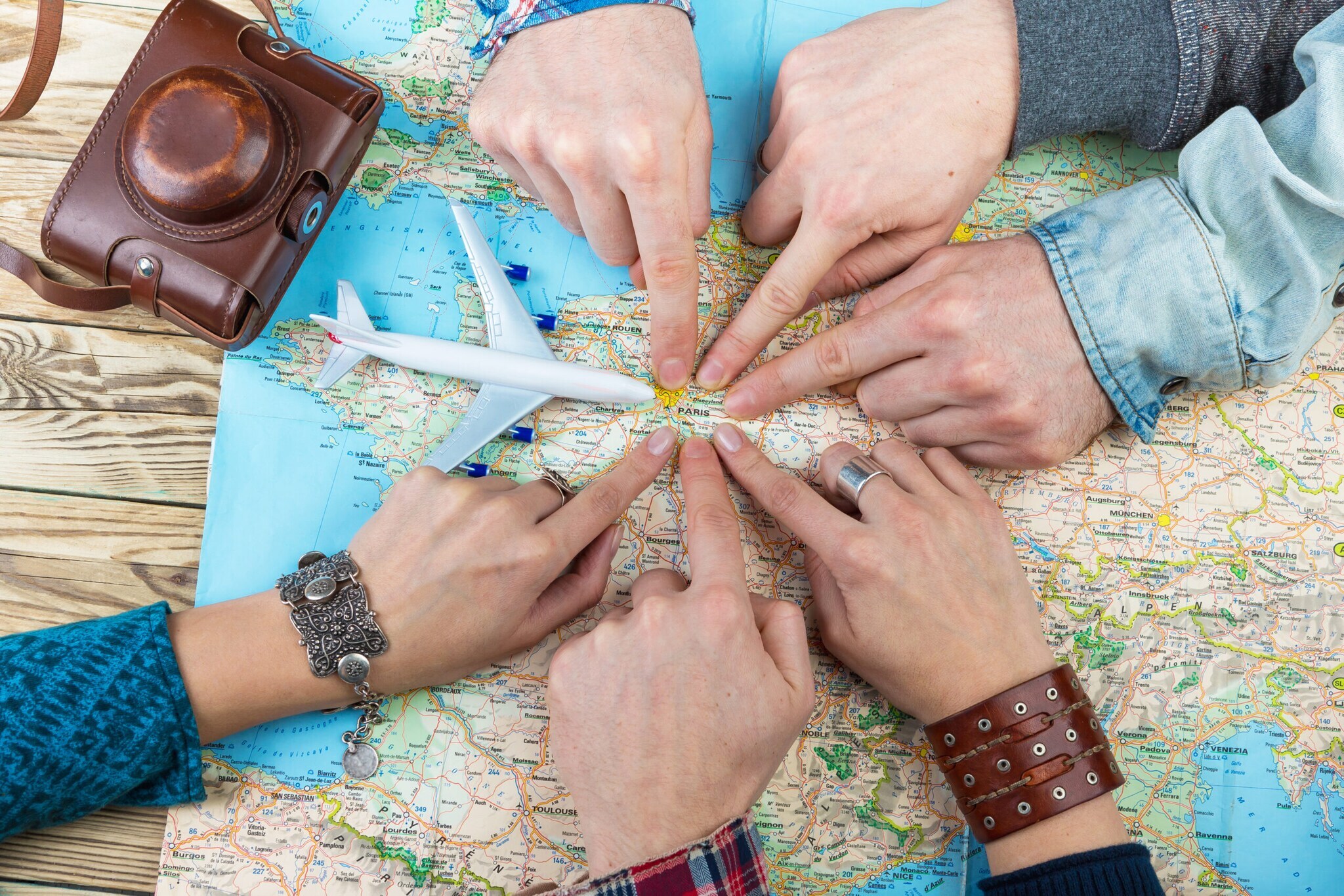 Sechs Personen, von denen nur die Hände zu sehen sind, zeigen auf einen Punkt auf einer Landkarte. Daneben liegen ein alter Fotoapparat und ein Modell eines Flugzeugs.