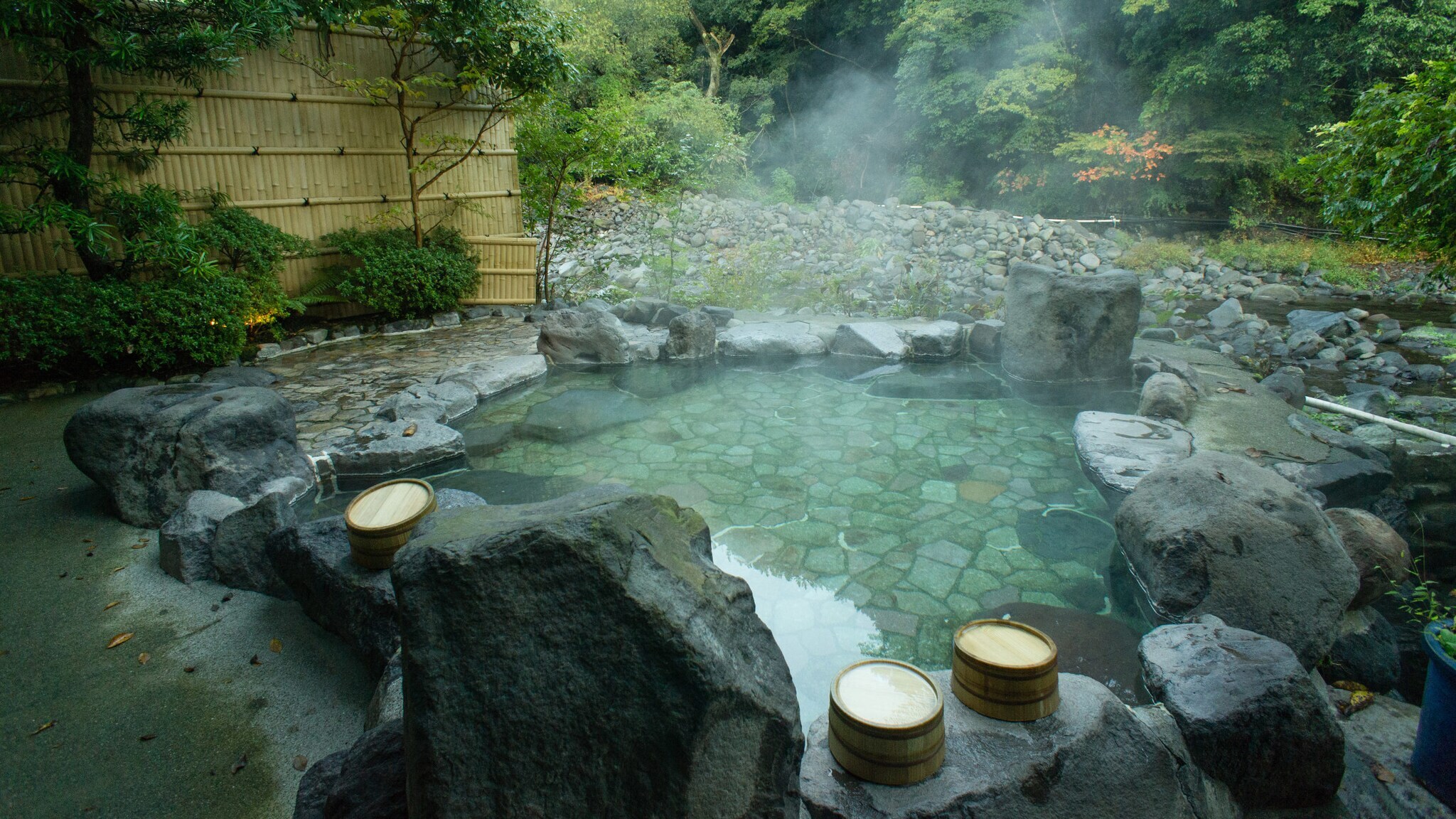 Flaches, dampfendes, von Steinen gerahmtes Thermalbad in einem japanischen Garten, im Vordergrund mehrere Saunakübel aus Holz auf Steinen.