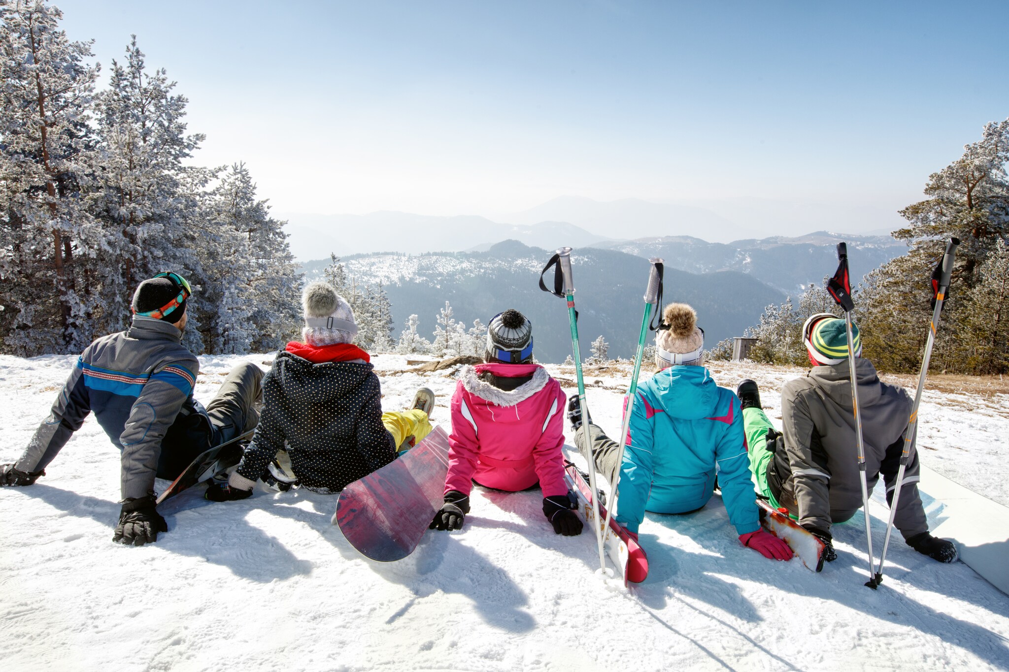 Fünf Personen mit Snowboards und Ski sitzen mit Blick auf eine weitere Berglandschaft auf einem Berg im Schnee.