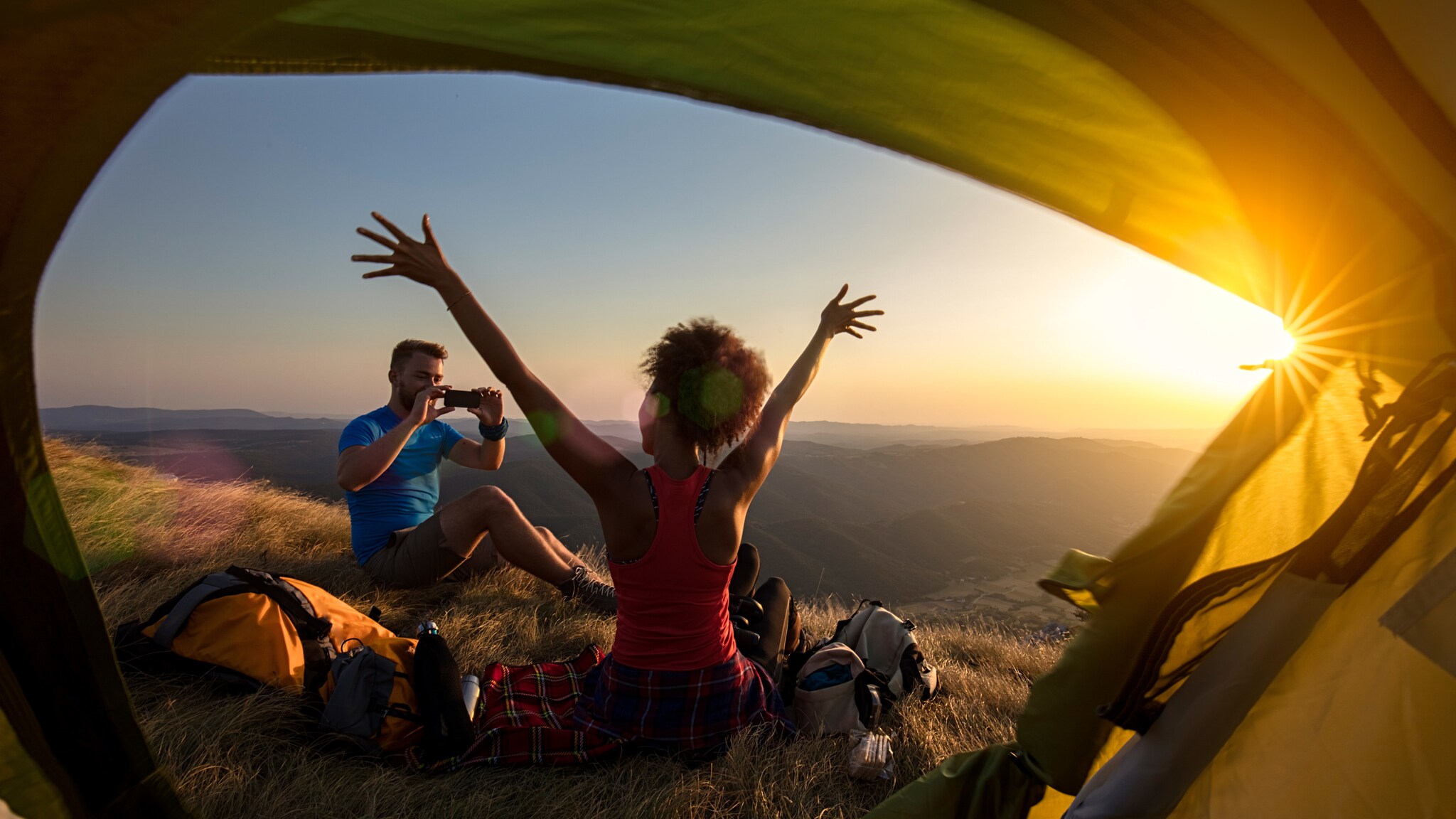 Blick aus einem Zelt auf zwei Personen auf einer Bergwiese bei Sonnenuntergang, eine Person macht ein Foto, die andere hebt die Arme in die Luft Blick aus einem Zelt auf zwei Personen auf einer Bergwiese bei Sonnenuntergang, eine Person macht ein Foto, die andere hebt die Arme in die Luft