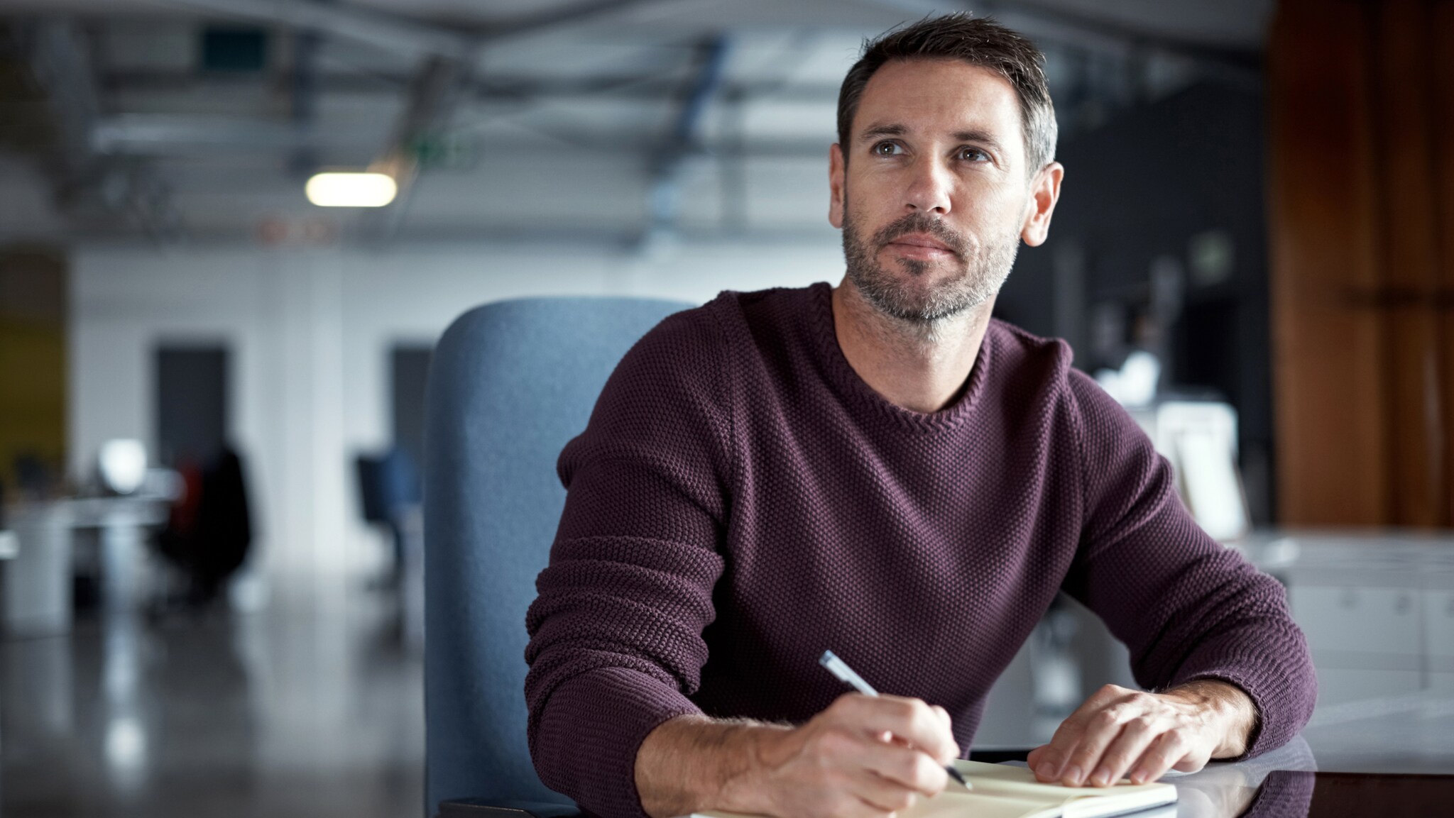 Mann mit braunem Pullover sitzt an Schreibtisch und schreibt in Notizbuch in modernem Büro