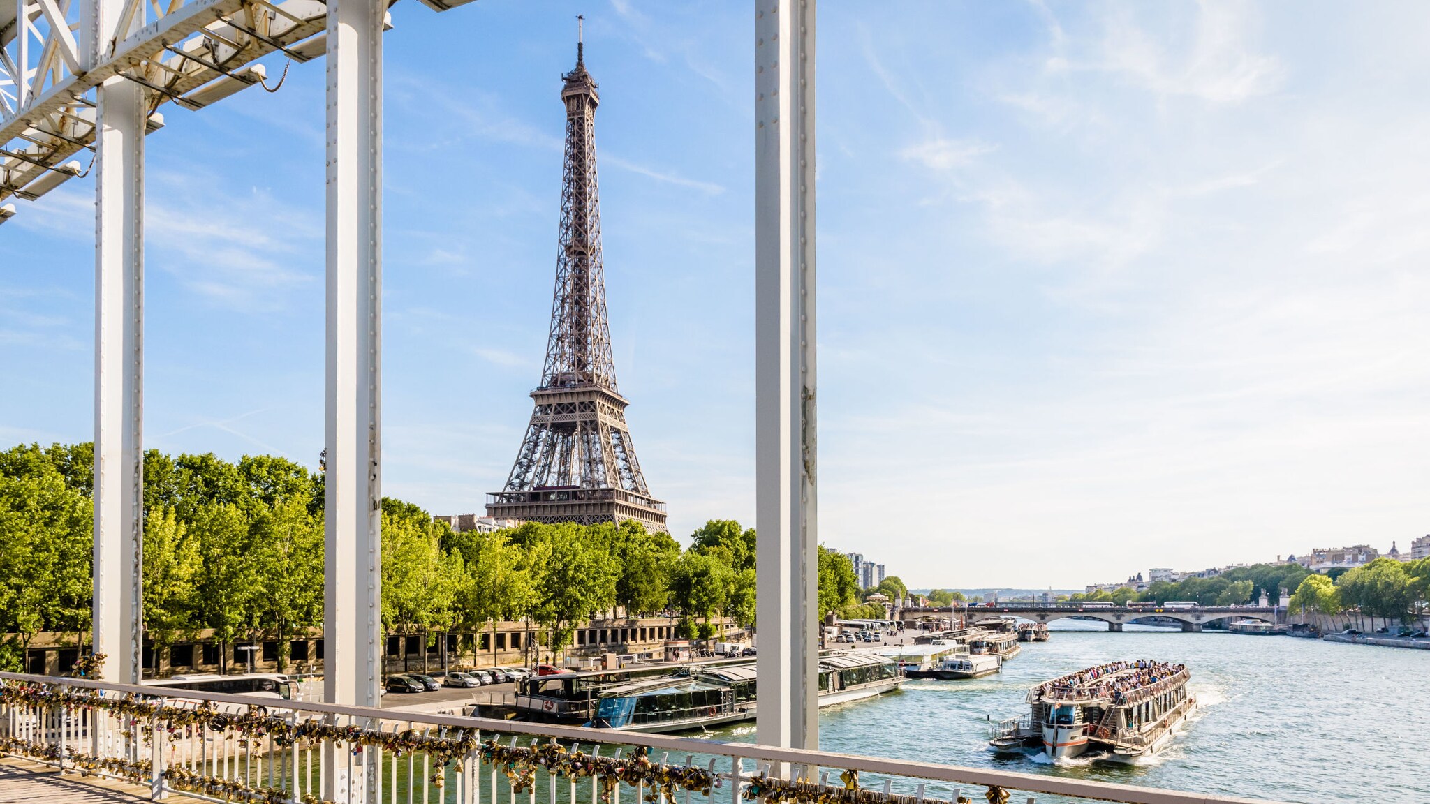 Eiffelturm hinter der Seine, gesehen von einer Brücke mit zahlreichen Liebesschlössern am Geländer, ein Ausflugsboot auf dem Fluss