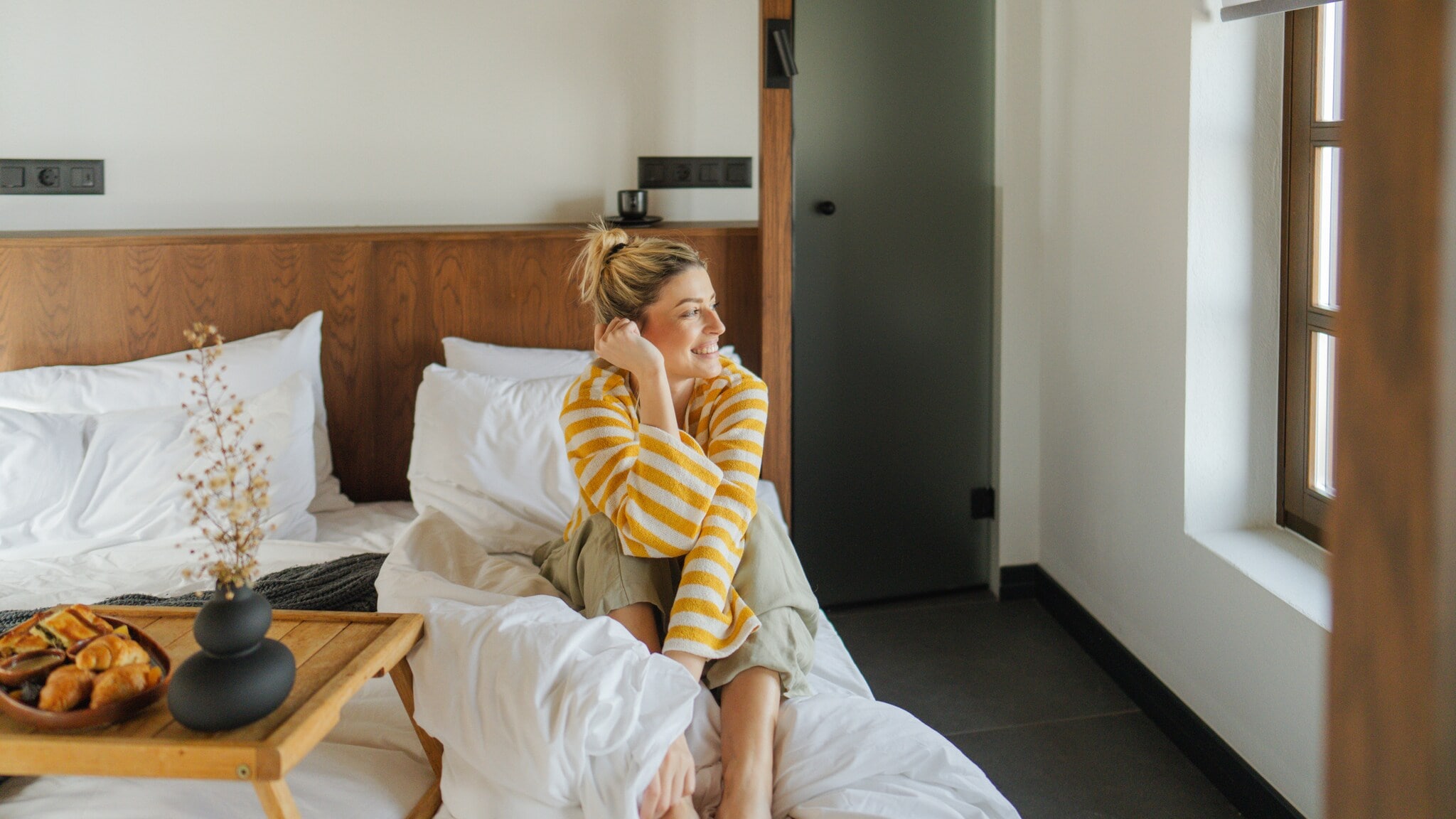 Person sitzt entspannt auf einem Bett mit weißer Bettwäsche in einem hellen Schlafzimmer mit Holzdetails und einem Fenster mit Jalousie Person sitzt entspannt auf einem Bett mit weißer Bettwäsche in einem hellen Schlafzimmer mit Holzdetails und einem Fenster mit Jalousie