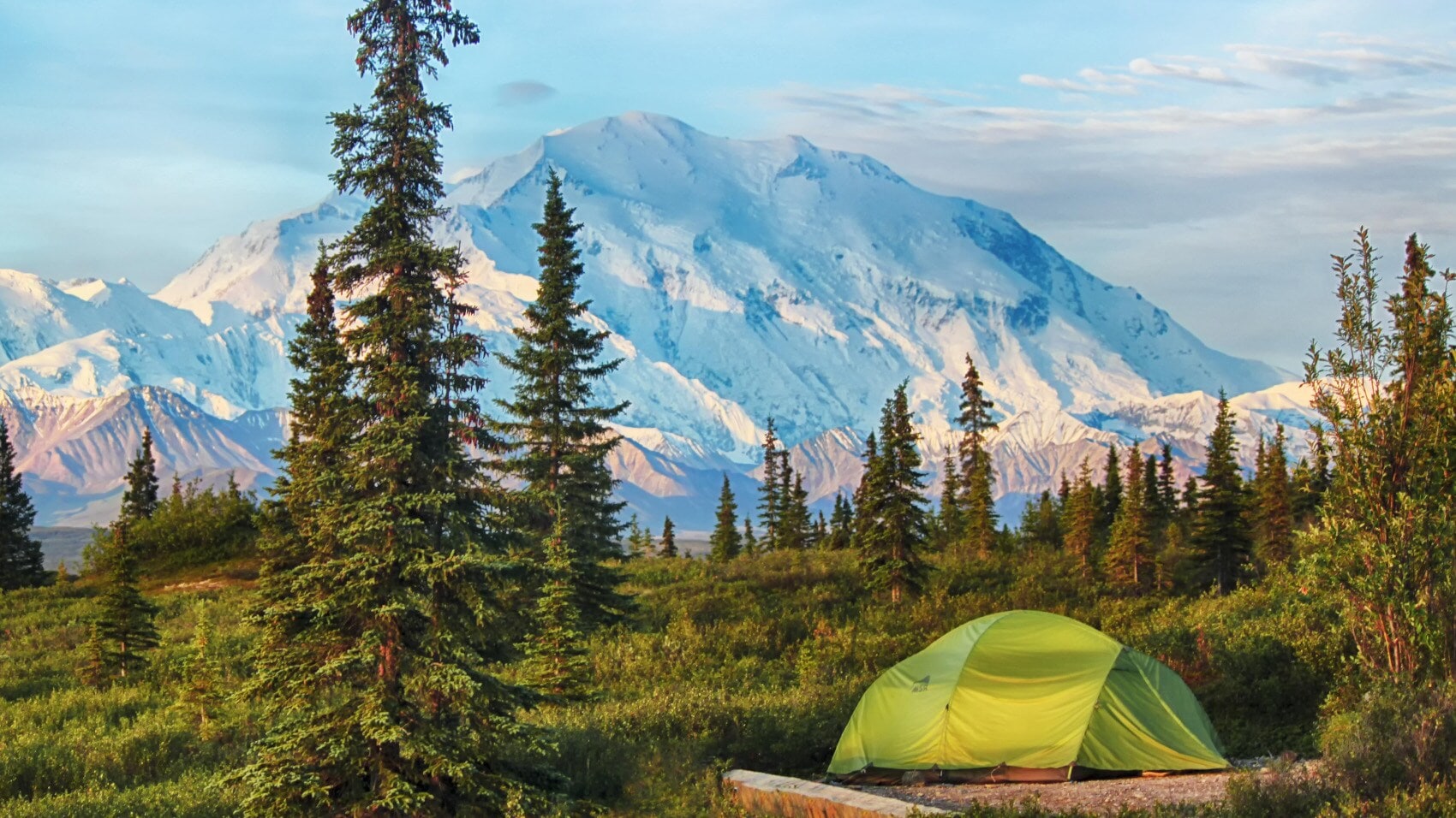 Grünes Zelt auf einer Wiese mit Nadelbäumen vor schneebedecktem Berg unter blauem Himmel Grünes Zelt auf einer Wiese mit Nadelbäumen vor schneebedecktem Berg unter blauem Himmel