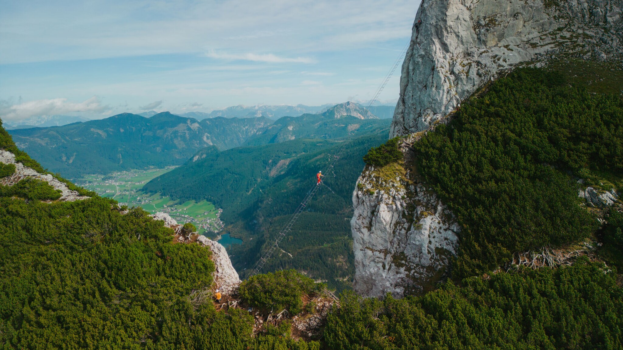 Eine Person auf einer Leiter, die hoch über einem Abgrund zu einem hohen Felsen führt, im Hintergrund Berge und Himmel.