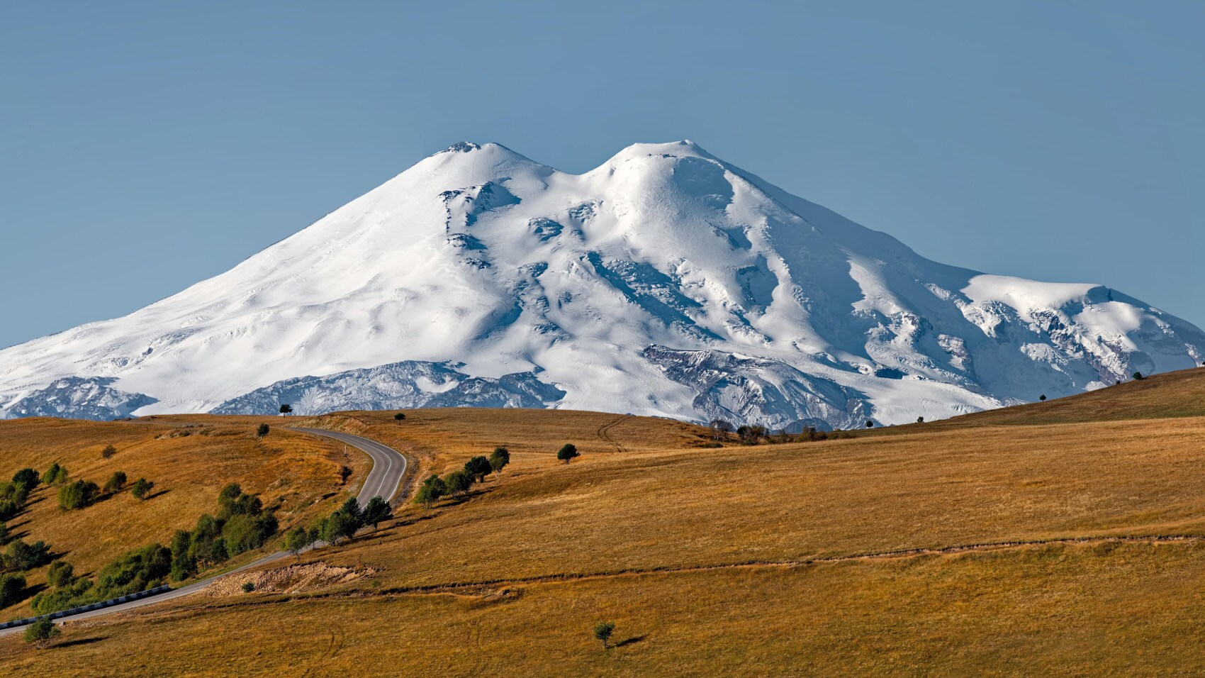 Schneebedeckter Berg mit zwei Gipfeln hinter hügeliger, brauner Landschaft und kurviger Straße unter klarem blauem Himmel