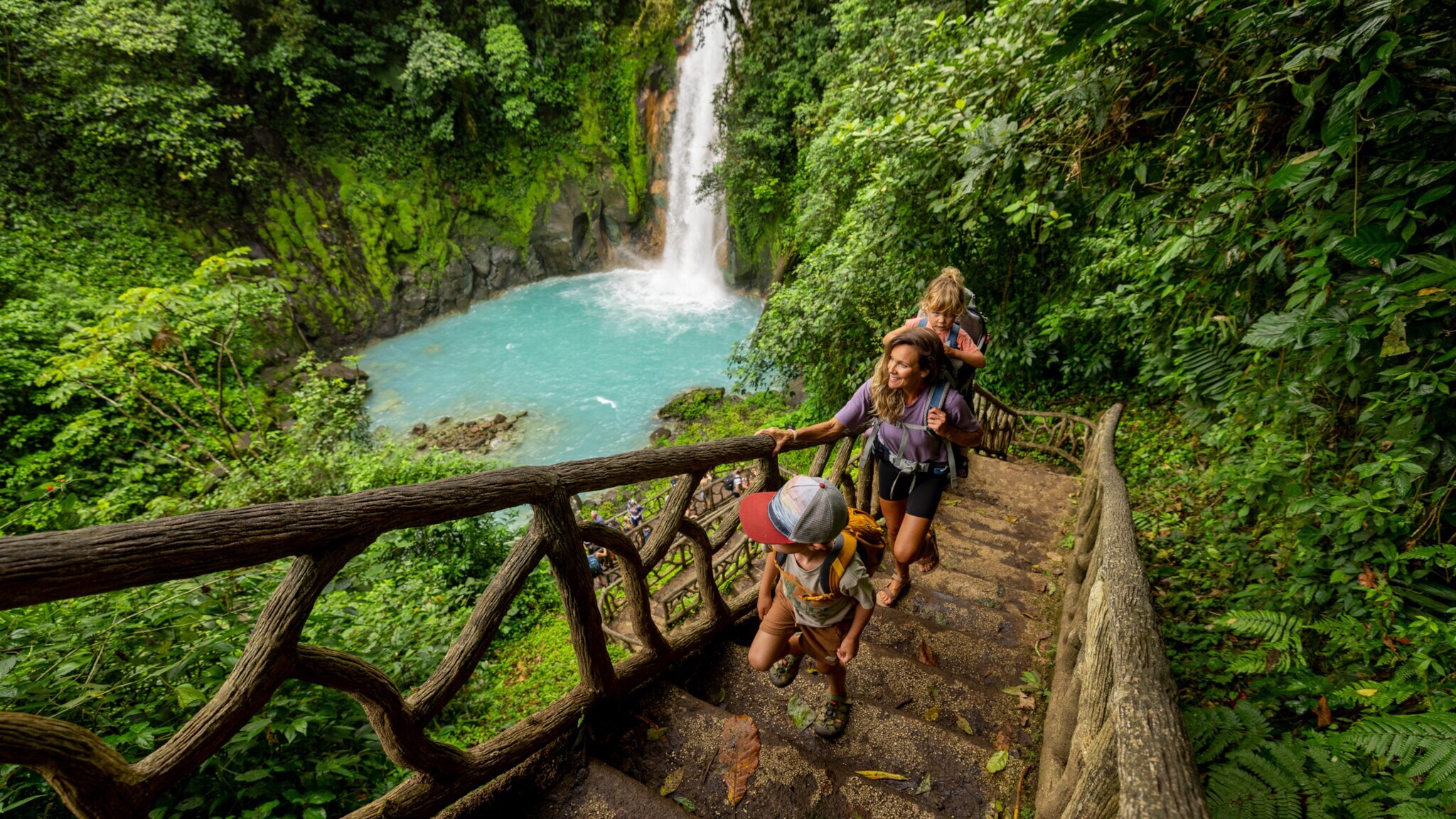 Drei Personen steigen eine Treppe aus Holz und Wurzeln hinauf, im Hintergrund ein Wasserfall und türkisfarbenes Wasser in einem dichten, grünen Dschungel.