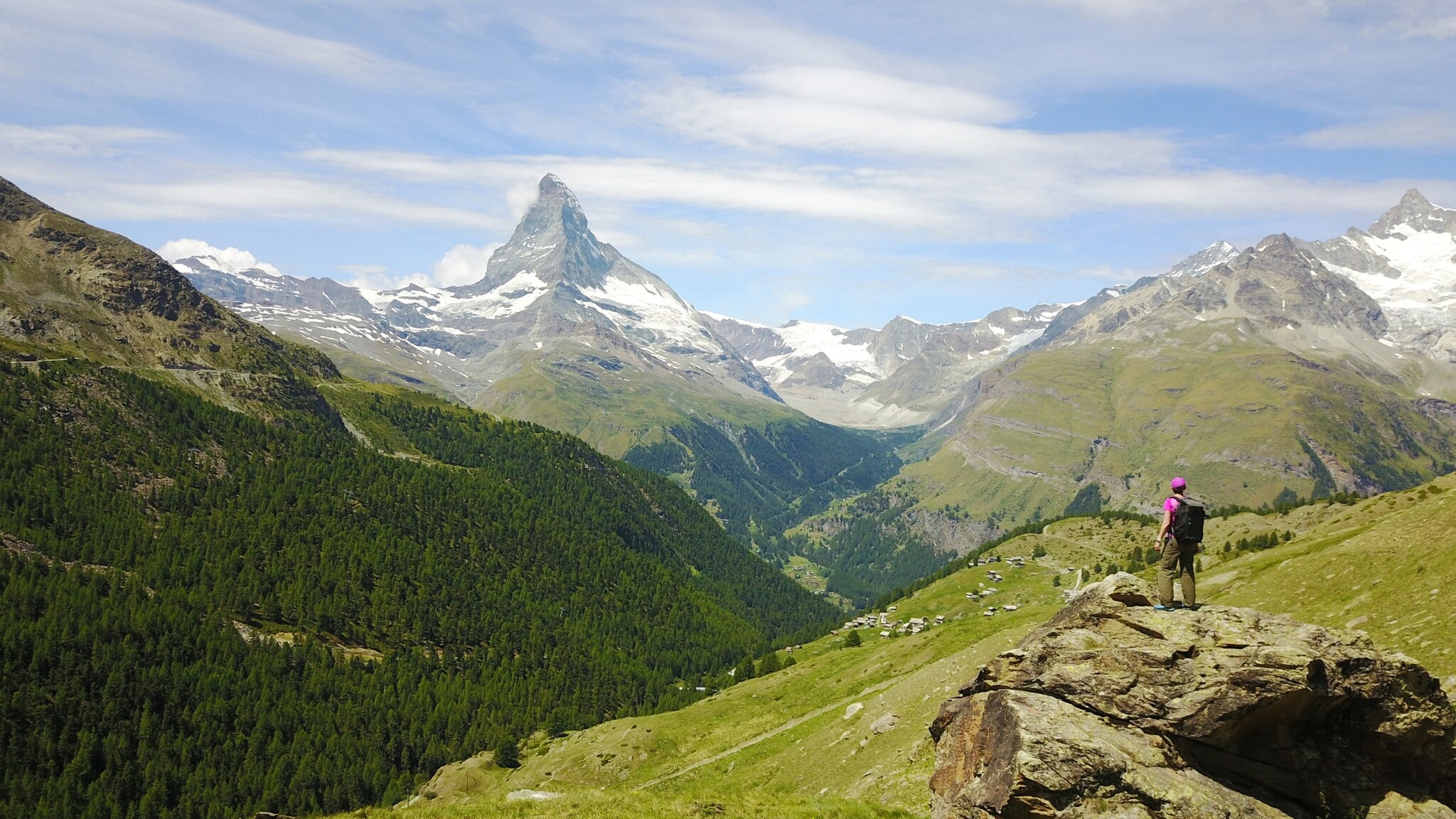Wanderer mit Rucksack steht auf Felsen und blickt auf das Matterhorn und grüne Alpenlandschaft Wanderer mit Rucksack steht auf Felsen und blickt auf das Matterhorn und grüne Alpenlandschaft