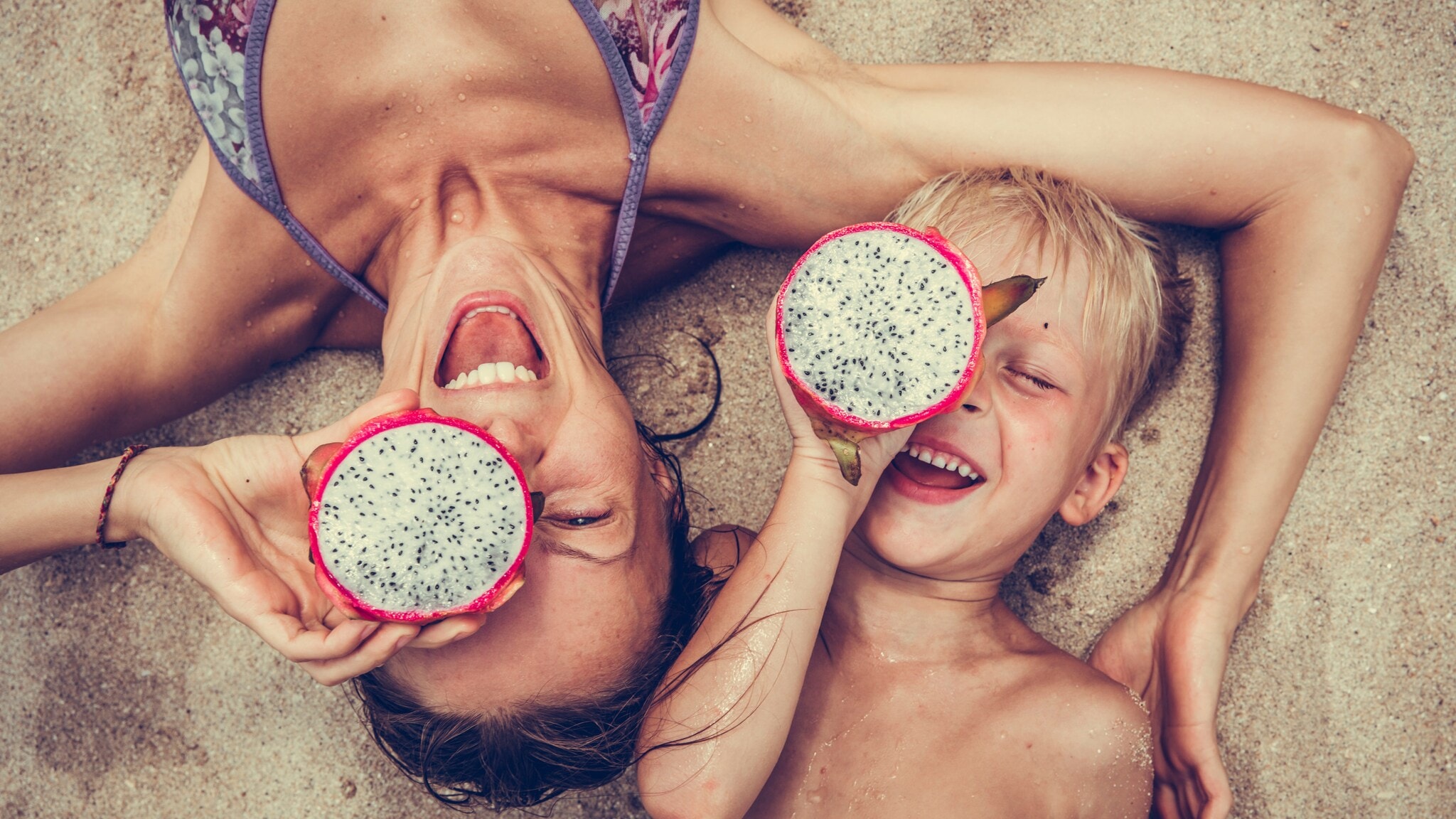 Eine Frau liegt mit einem Kind im Sand, beide halten eine tropische Frucht vor einem ihrer Augen. Eine Frau liegt mit einem Kind im Sand, beide halten eine tropische Frucht vor einem ihrer Augen.