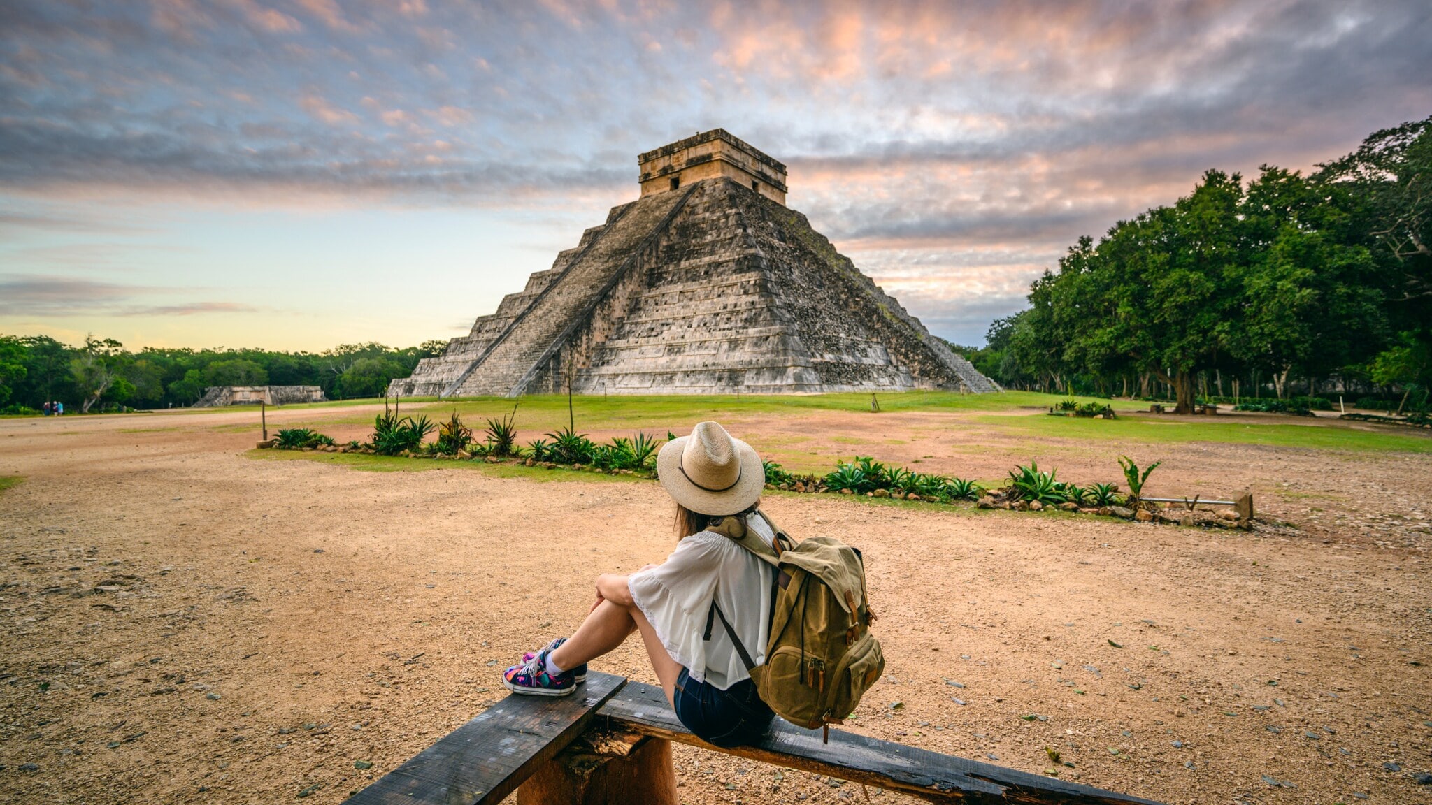 Person mit Hut und Rucksack sitzt auf einer Bank und blickt auf die Pyramide von Chichén Itzá bei Sonnenuntergang Person mit Hut und Rucksack sitzt auf einer Bank und blickt auf die Pyramide von Chichén Itzá bei Sonnenuntergang