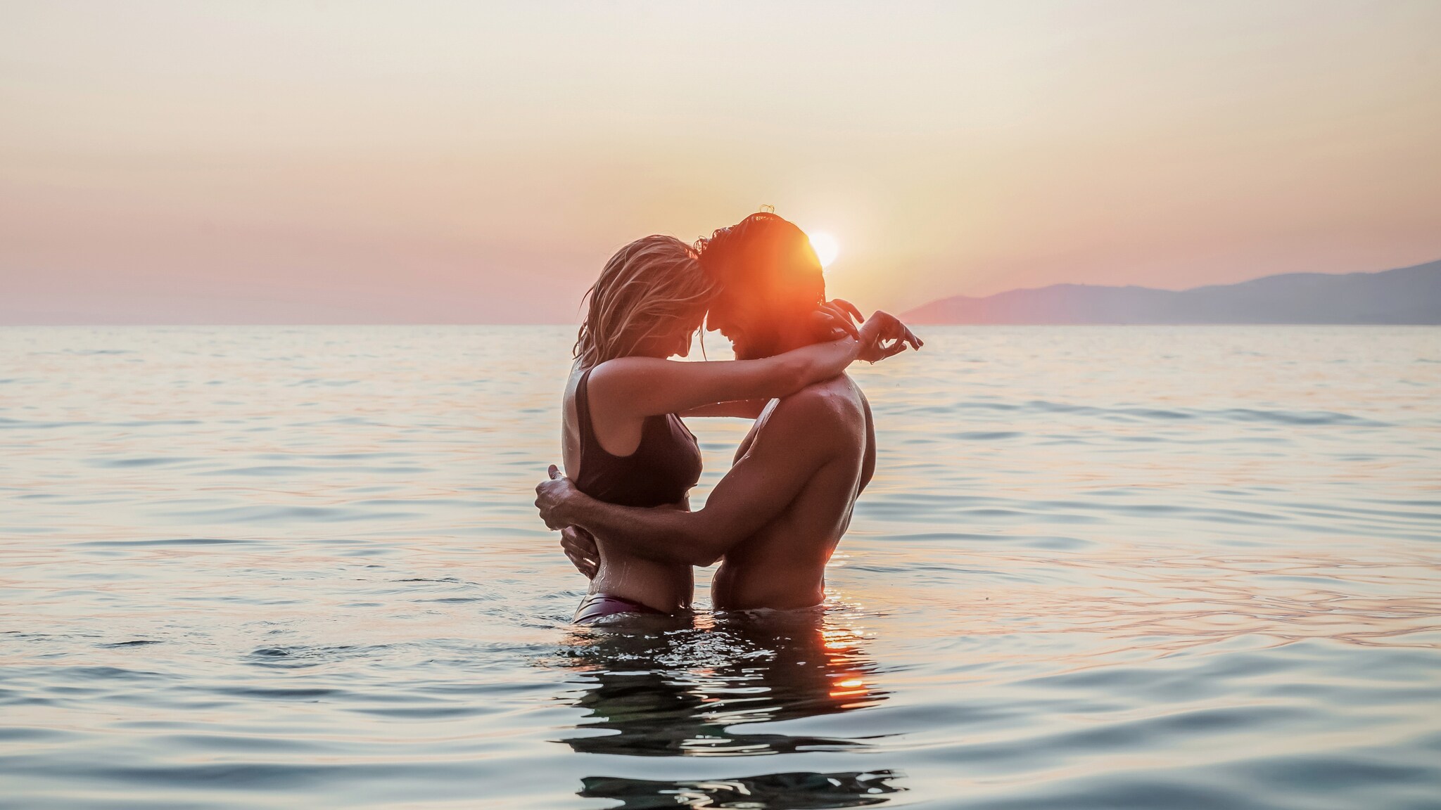 Paar umarmt sich im Meer bei Sonnenuntergang, Wasser bis zur Taille, sanfte Wellen und Berge im Hintergrund