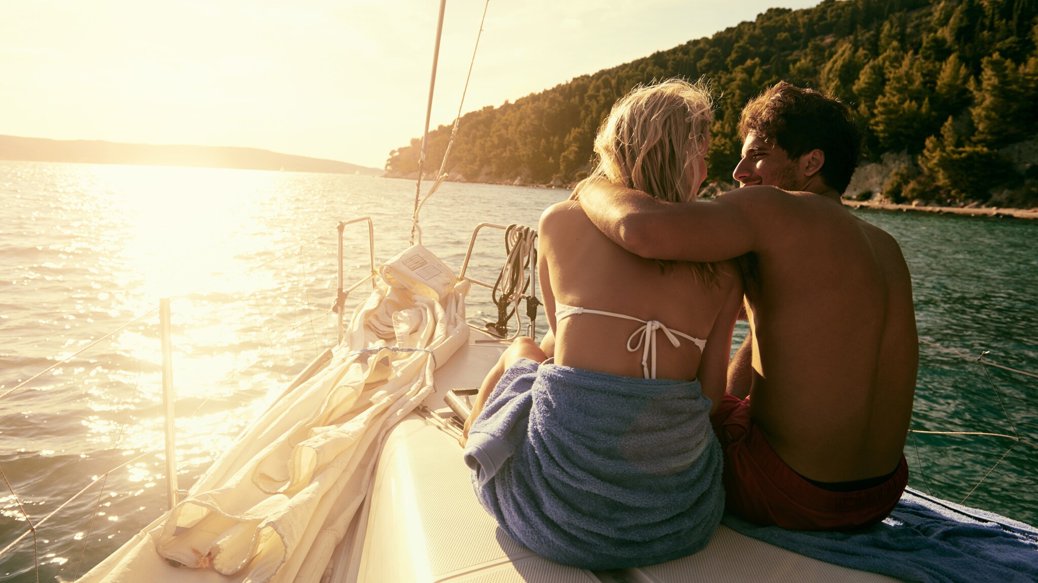 Paar sitzt eng umschlungen auf einem Boot bei Sonnenuntergang, umgeben von Wasser und bewaldeten Hügeln