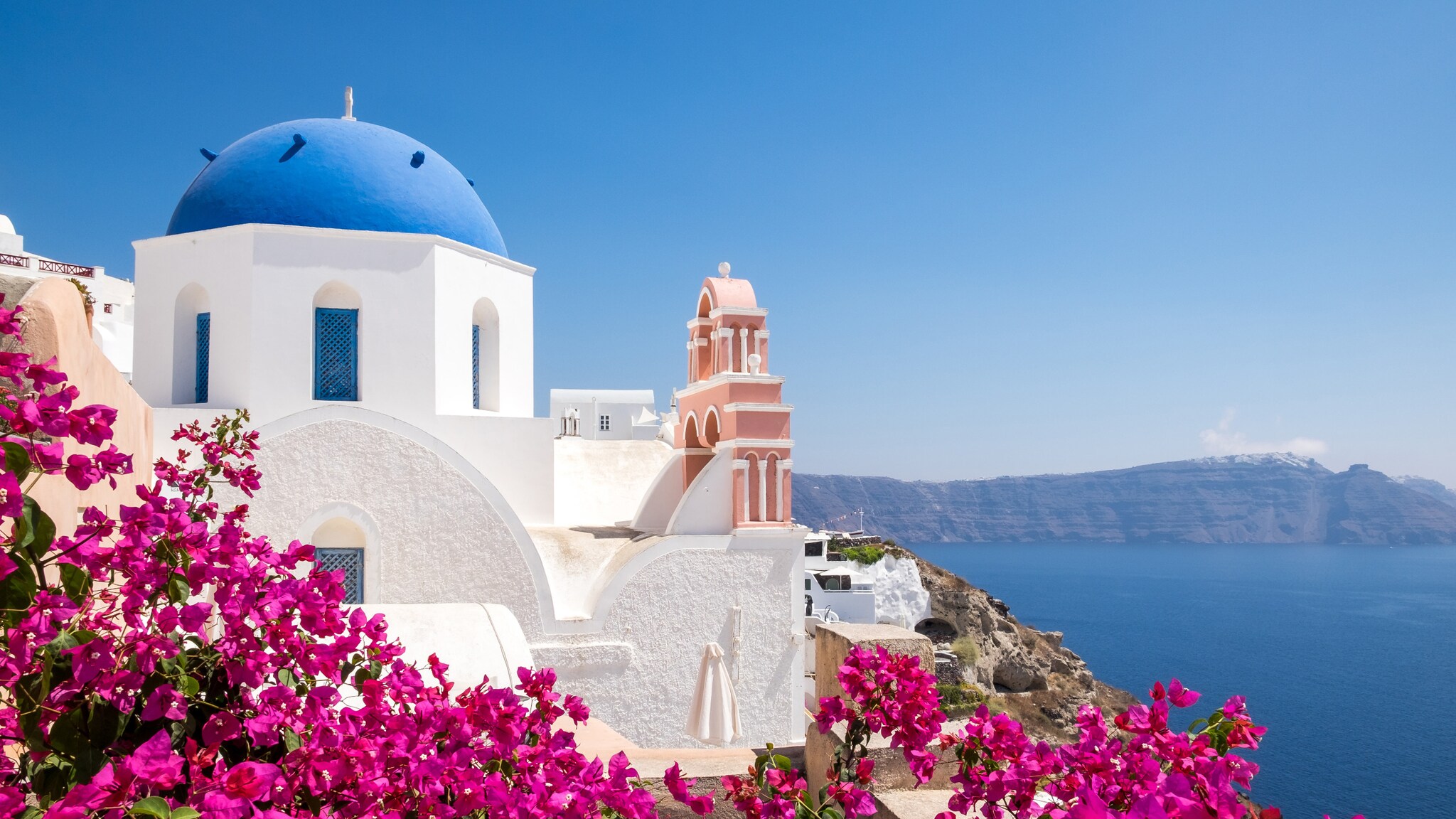 Blick auf eine weiße Kirche mit blauem Kuppeldach und pinken Bougainvillea-Blüten in Santorin, Griechenland, mit Meer und Klippen im Hintergrund