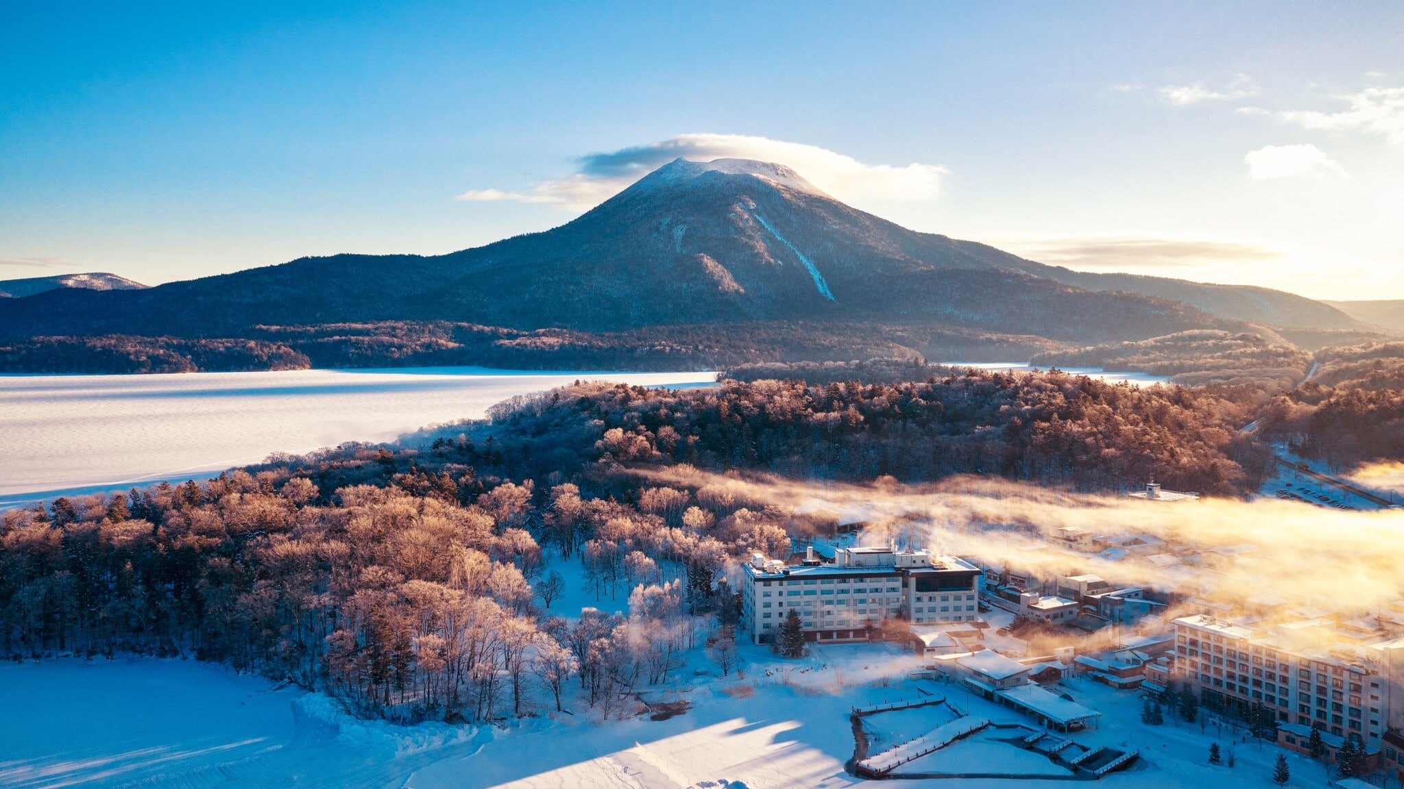 Verschneite Landschaft mit Wald und Gebäuden vor einem großen Berg unter klarem Himmel bei Sonnenaufgang Verschneite Landschaft mit Wald und Gebäuden vor einem großen Berg unter klarem Himmel bei Sonnenaufgang