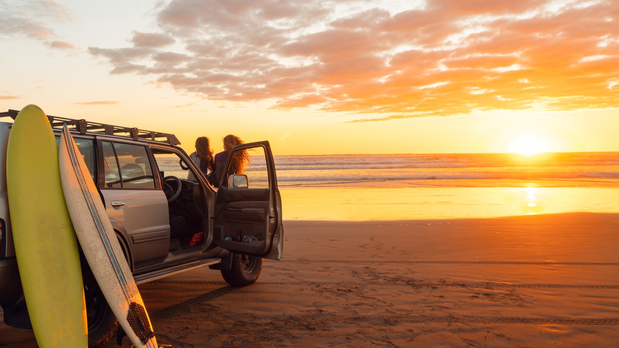 Zwei Personen stehen neben einem offenen Auto am Strand mit zwei Surfbrettern, Sonnenuntergang über dem Meer im Hintergrund