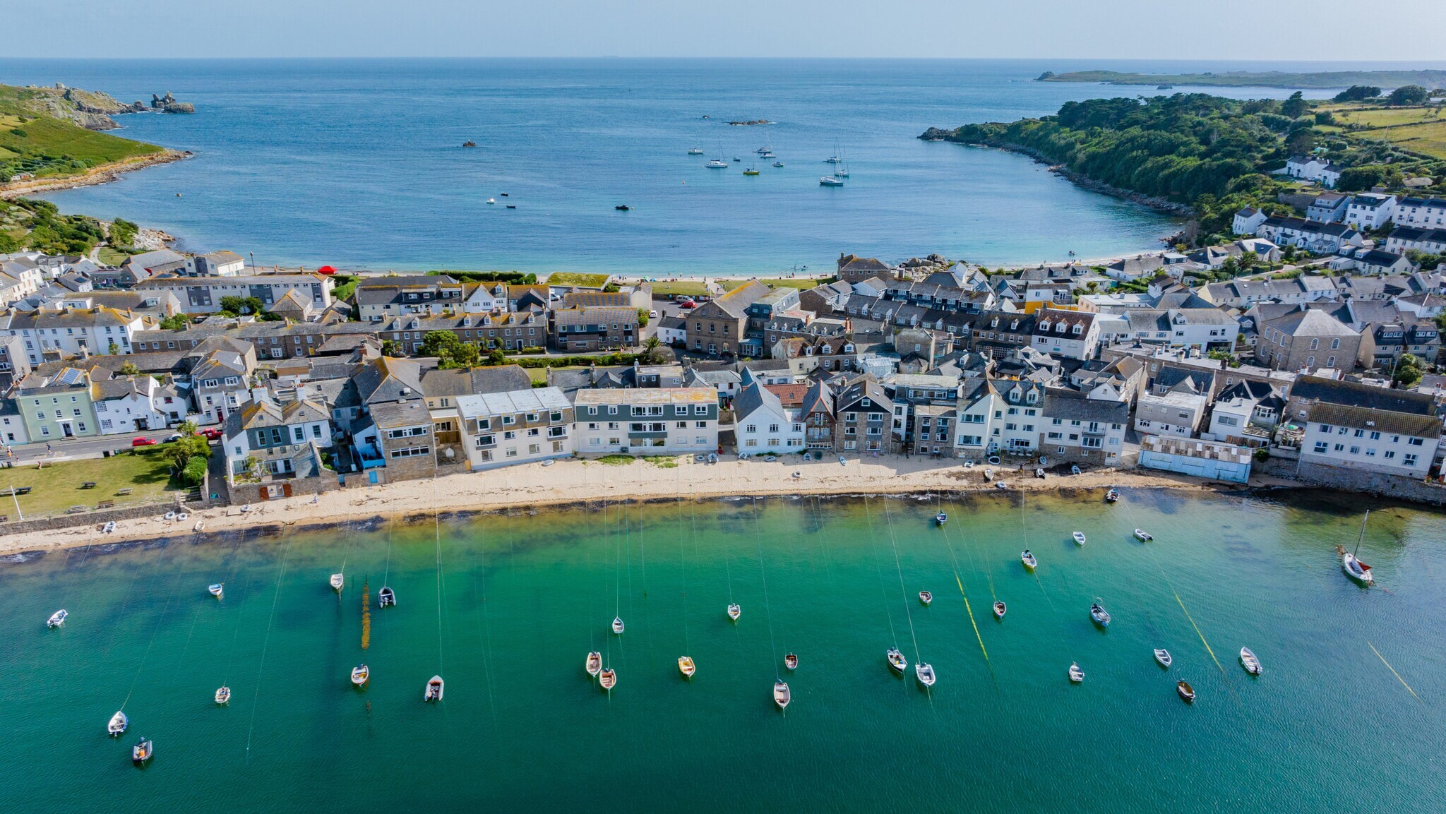 Luftaufnahme eines Küstendorfes mit weißen Häusern, einem Sandstrand und zahlreichen Booten im klaren, grünen Wasser einer Bucht