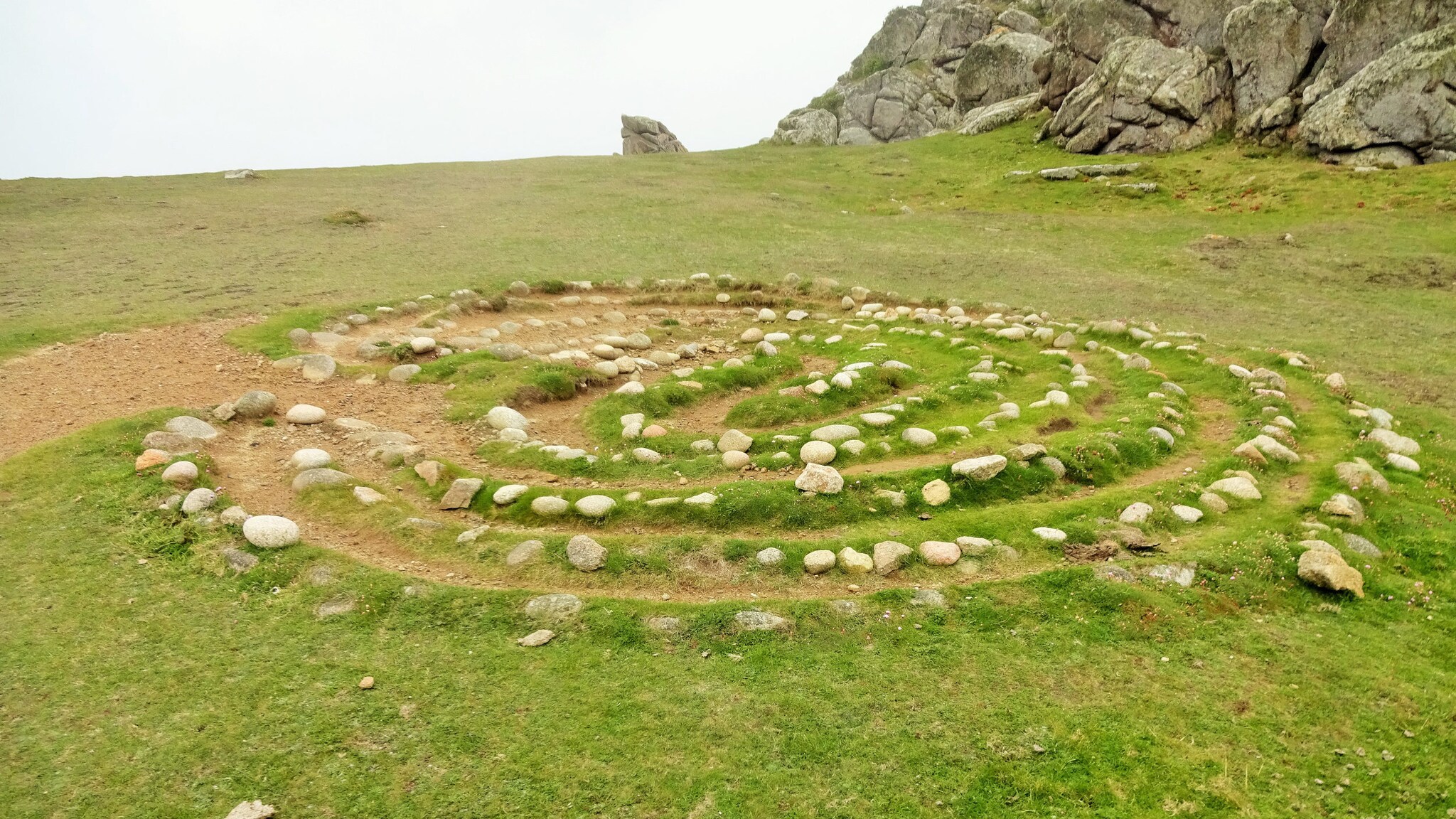 Steinspirale auf einer grasbewachsenen Fläche vor Felsen unter bewölktem Himmel