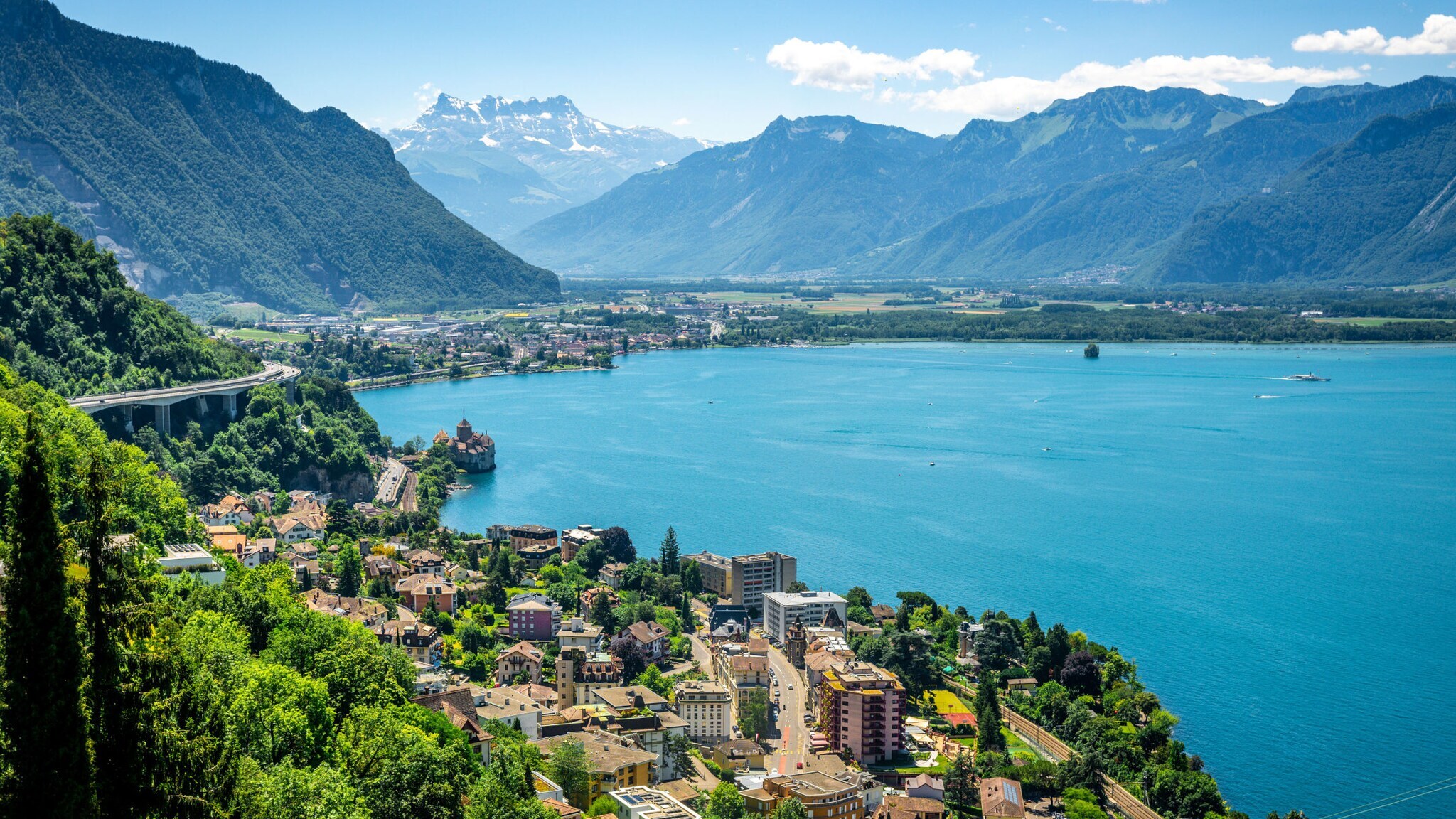 Blick auf den Genfersee mit der Stadt Montreux, umgeben von grünen Bergen und schneebedeckten Alpen im Hintergrund Blick auf den Genfersee mit der Stadt Montreux, umgeben von grünen Bergen und schneebedeckten Alpen im Hintergrund