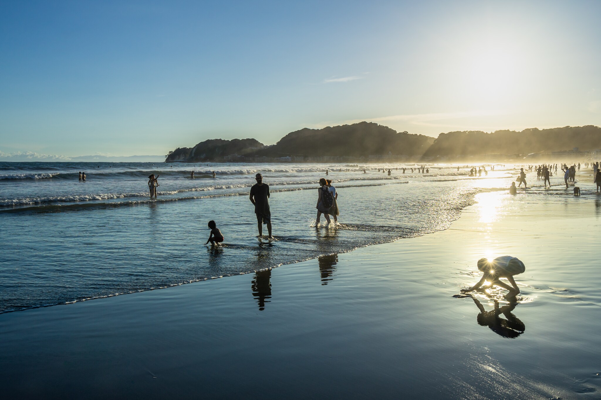 Silhouetten zahlreicher Personen im flachen Wasser an einem Sandstrand bei Gegenlicht.