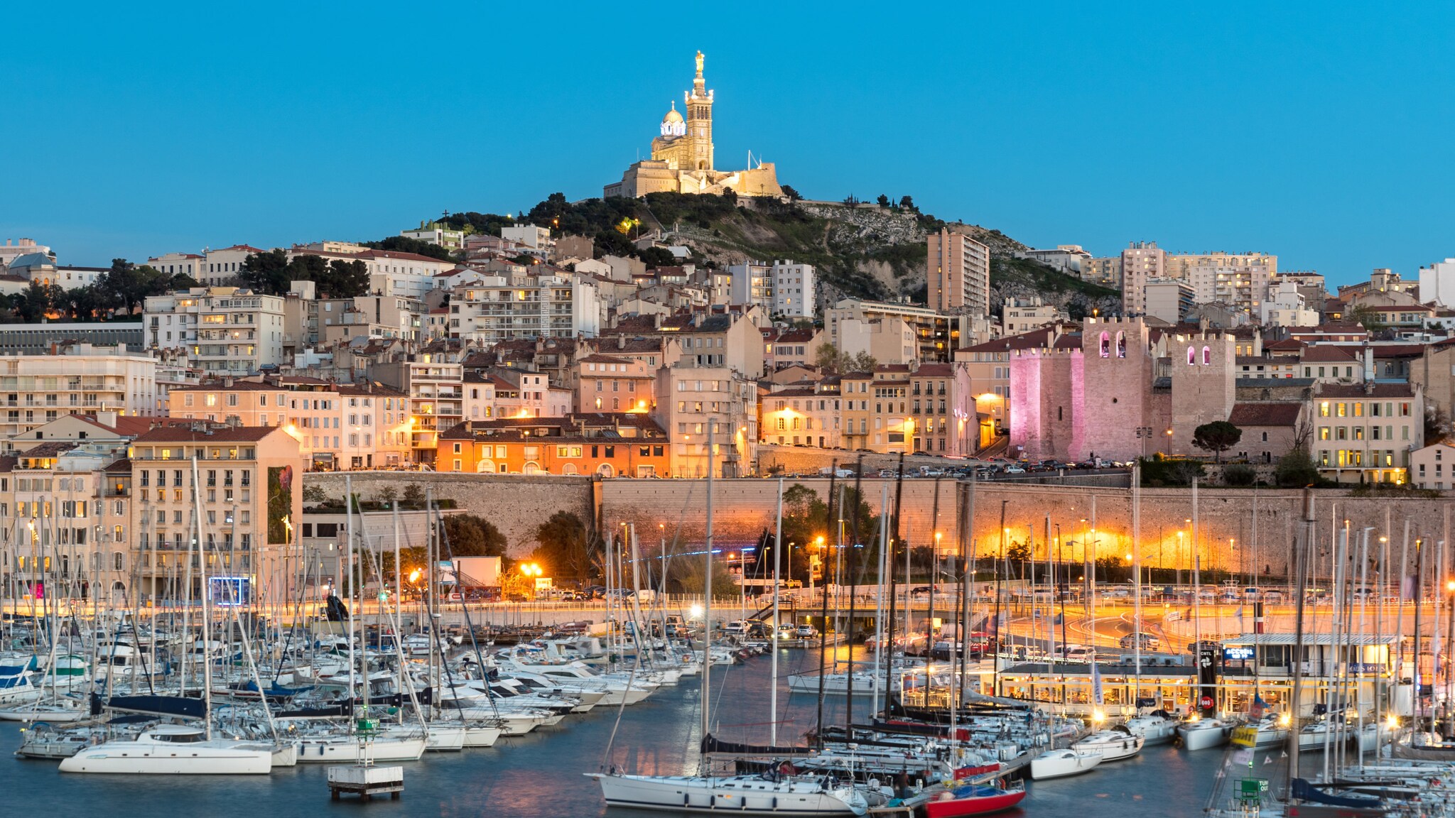 Abendliche Hafenansicht von Marseille mit Segelbooten im Wasser und der beleuchteten Kirche Notre-Dame de la Garde auf einem Hügel im Hintergrund