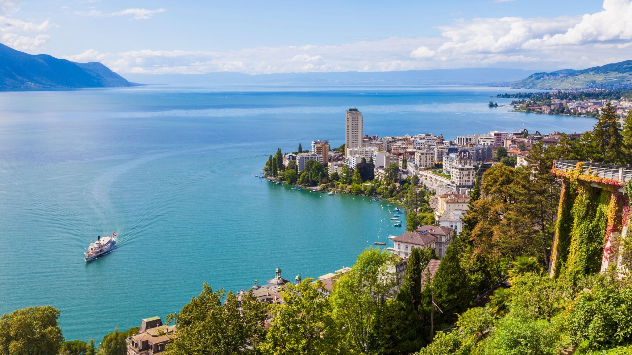Blick auf den Genfersee mit einem Ausflugsboot, umgeben von Bergen und der Stadt Montreux am Ufer Blick auf den Genfersee mit einem Ausflugsboot, umgeben von Bergen und der Stadt Montreux am Ufer