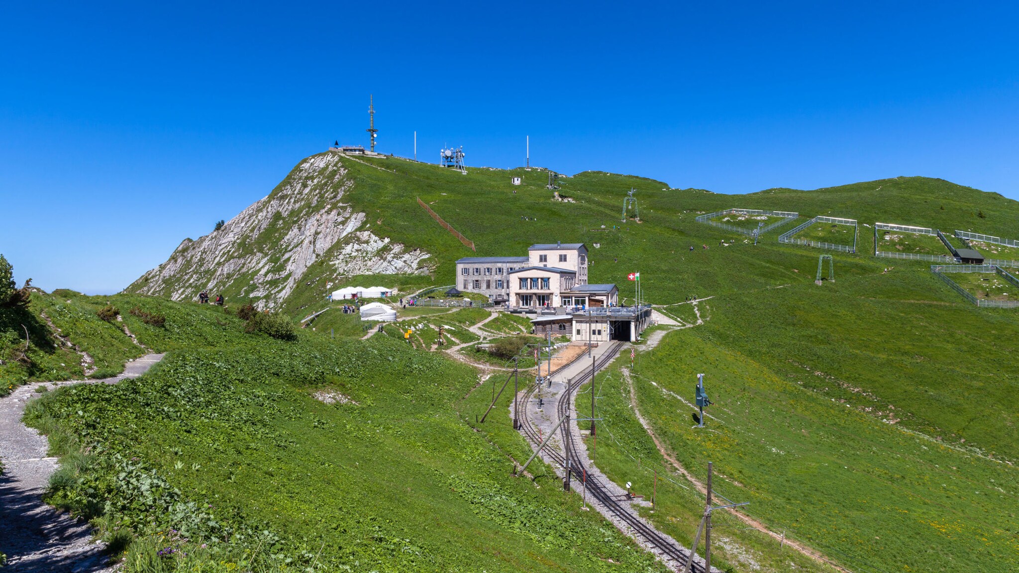 Grüner Berg mit Wanderweg, Bergbahn und Gebäude unter klarem blauem Himmel