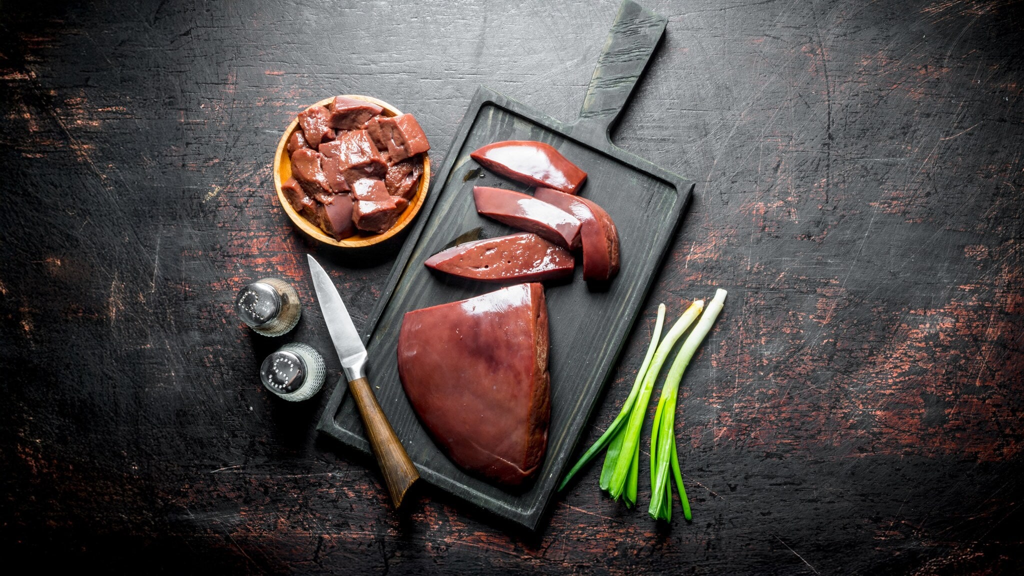 Rohe Leberstücke auf schwarzem Schneidebrett mit Messer, Frühlingszwiebeln und Gewürzstreuern auf dunklem Untergrund Rohe Leberstücke auf schwarzem Schneidebrett mit Messer, Frühlingszwiebeln und Gewürzstreuern auf dunklem Untergrund