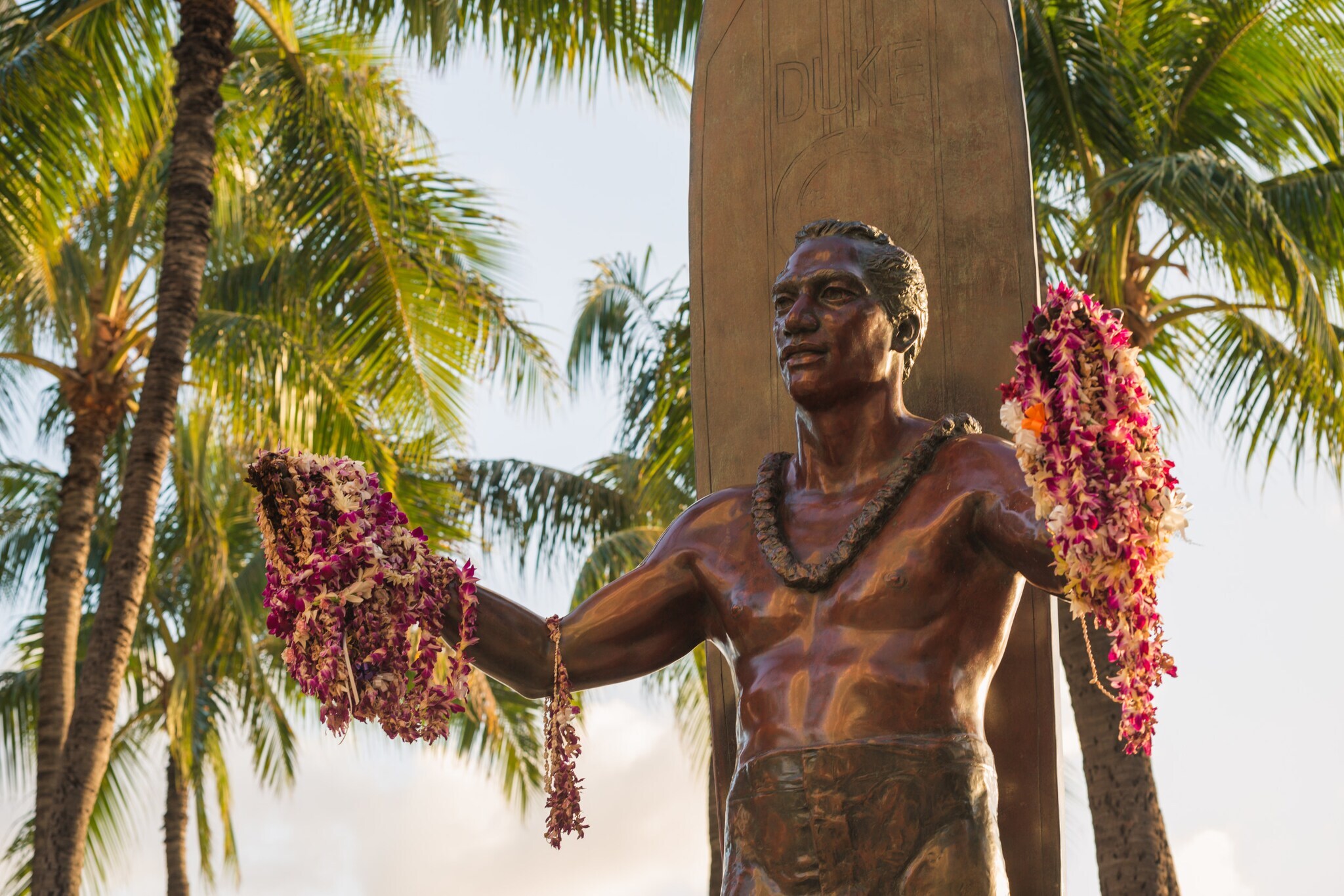 Bronzestatue von Surf-Legende Duke Kahanamoku mit ausgebreiteten Armen, an denen Blumenketten hängen.