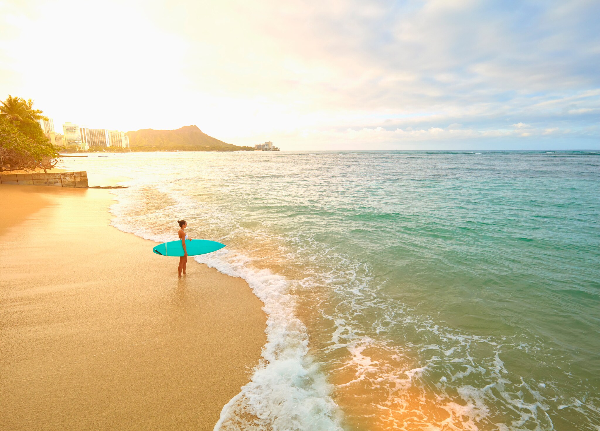 Eine Frau mit türkisfarbenem Surfboard unterm Arm steht am frühen Morgen am leeren Strand von Waikiki.