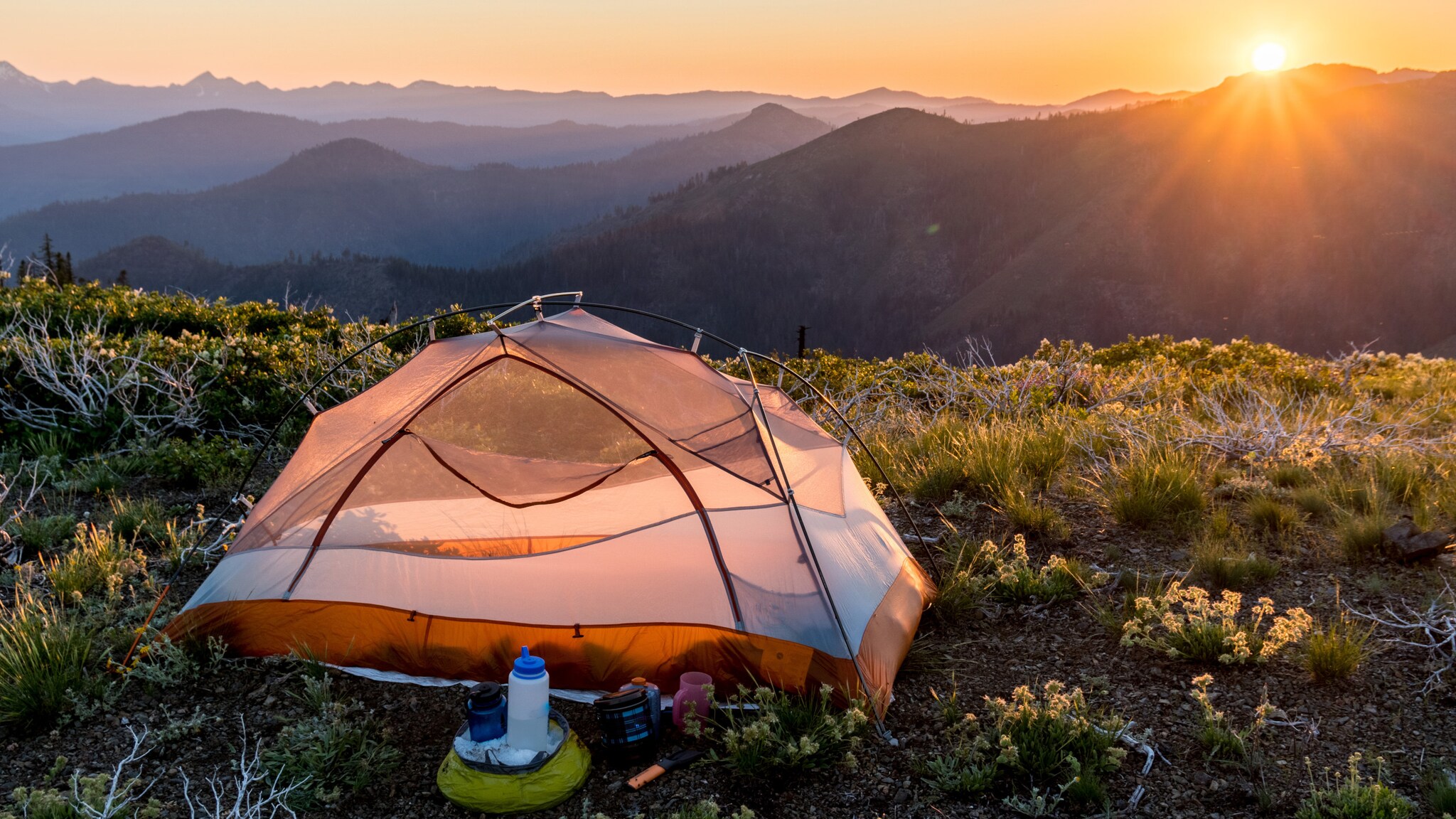 Zelt auf Berggipfel bei Sonnenaufgang mit Blick auf mehrere Bergketten und Campingausrüstung im Vordergrund