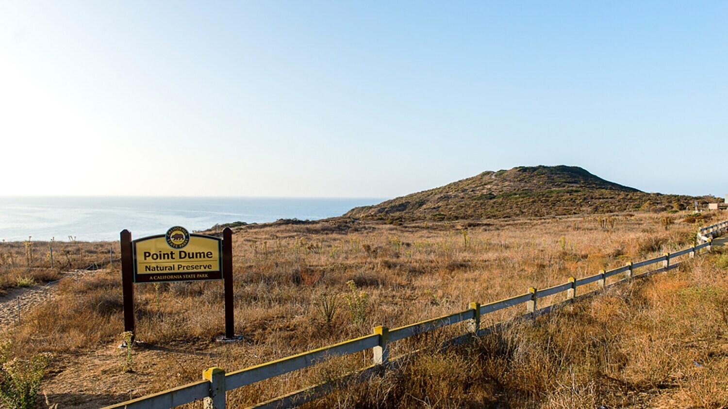 Schild mit der Aufschrift Point Dume Natural Preserve vor trockenem Grasland und einem Hügel, im Hintergrund das Meer.