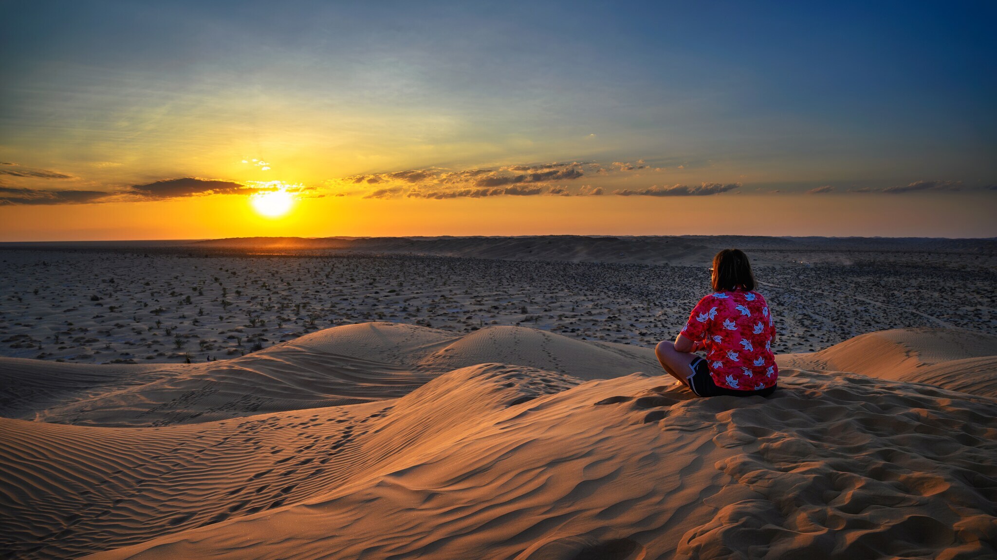 Person in rotem Hemd mit weißen Mustern sitzt auf Sanddüne und blickt auf Sonnenuntergang über Wüste